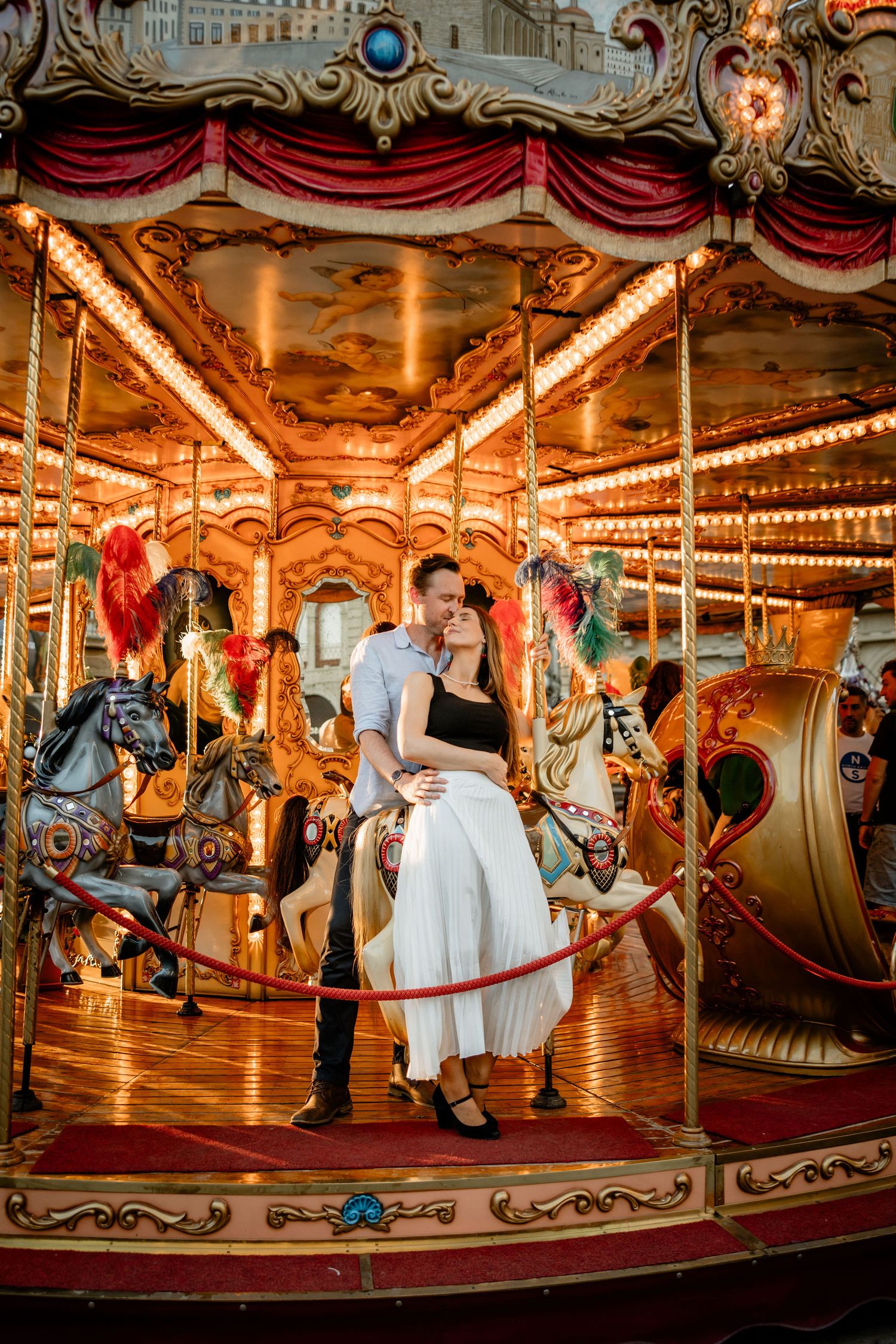 Romantic couple poses together on a vintage carousel with ornate painted horses glowing under warm lights.