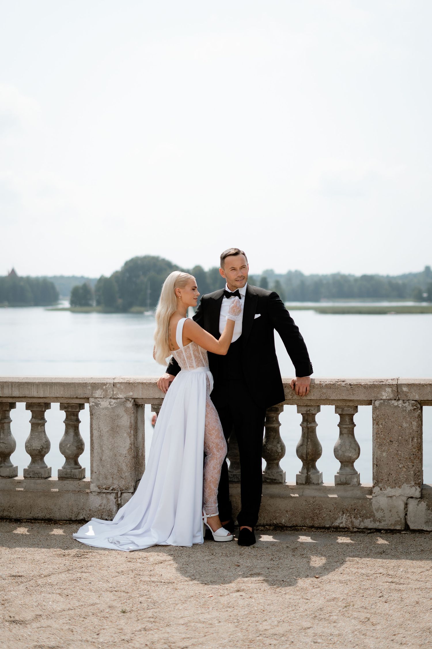 Elegant couple in wedding attire poses against stone balustrade with lake view.