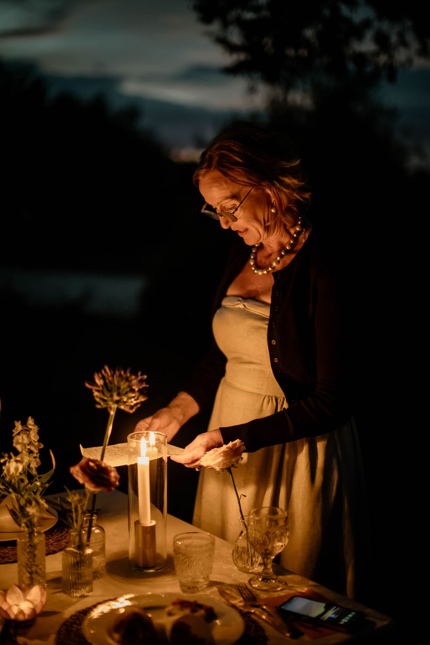 A person illuminated by candlelight tends to an elegant table setting at dusk.