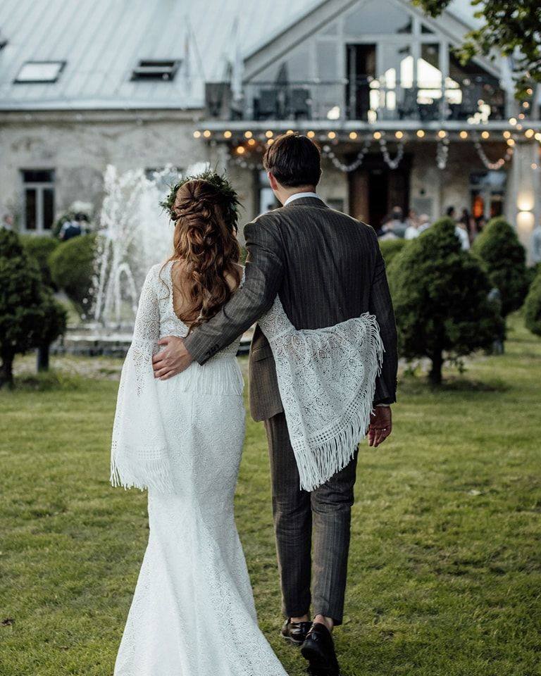 A couple in wedding attire walking away together on a grassy lawn with buildings in the background.
