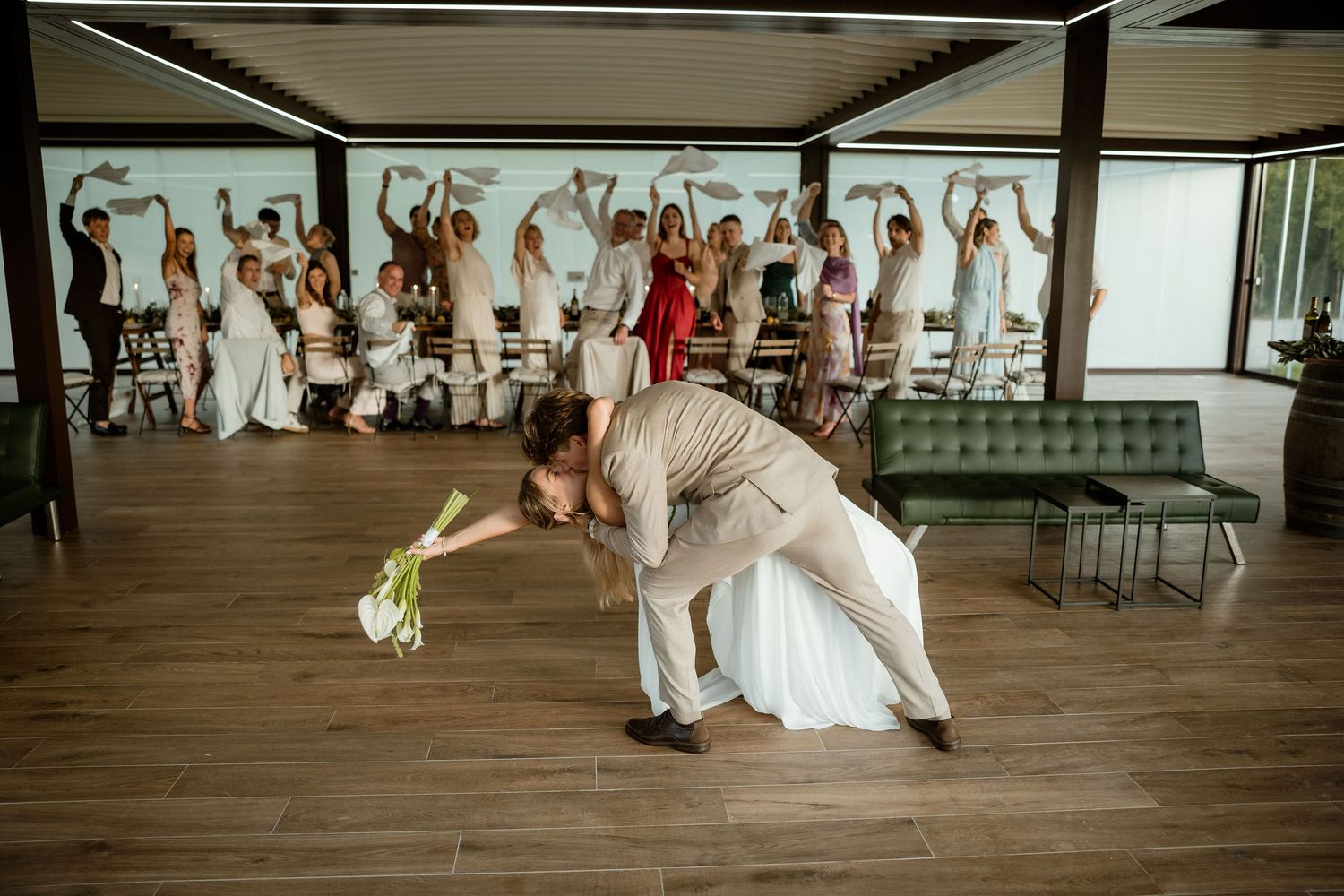 A couple performs a dramatic dance dip while wedding guests cheer and raise their hands in celebration on a wooden dance floor.