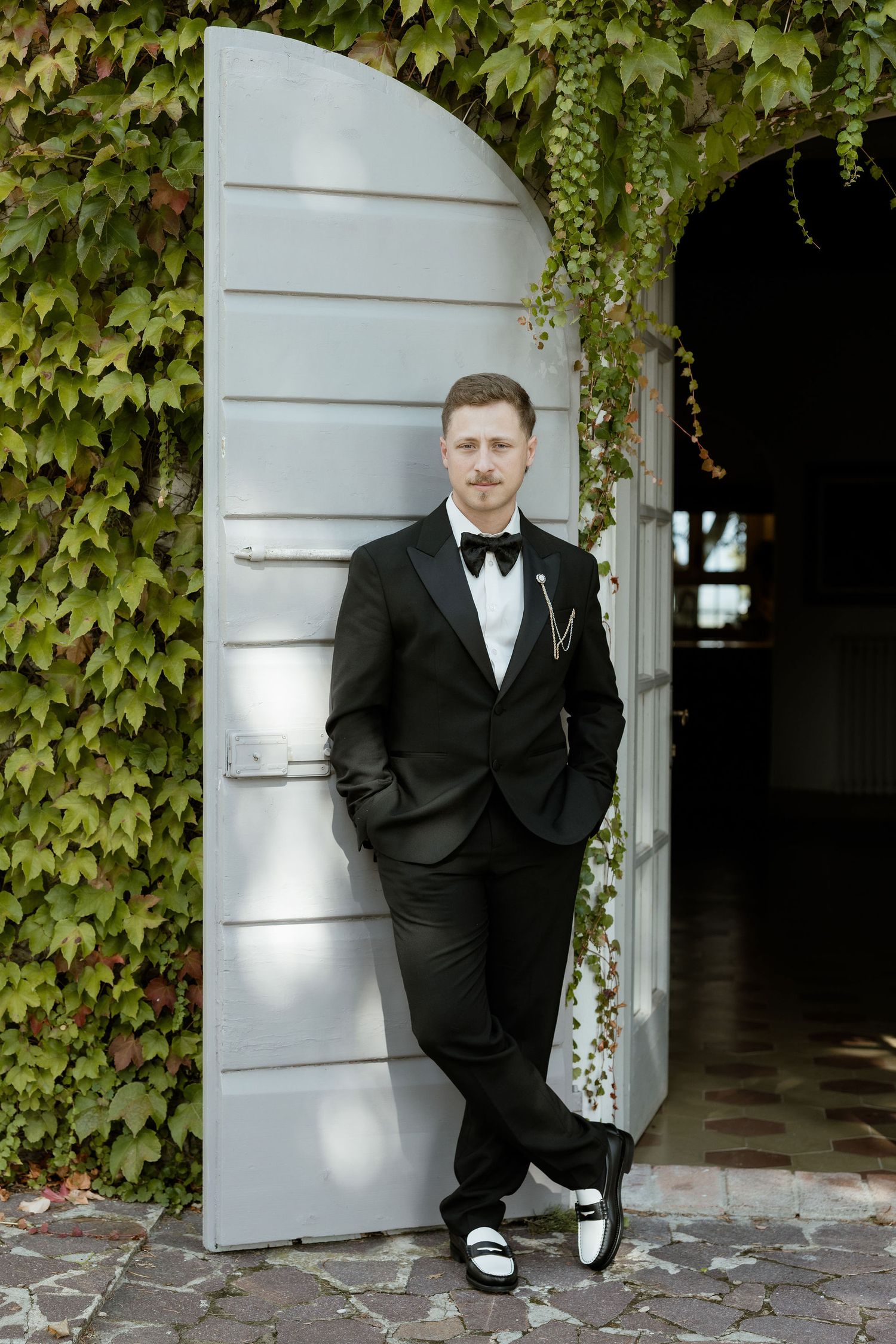 A person in a black tuxedo poses against a white column with ivy-covered walls in a formal setting.