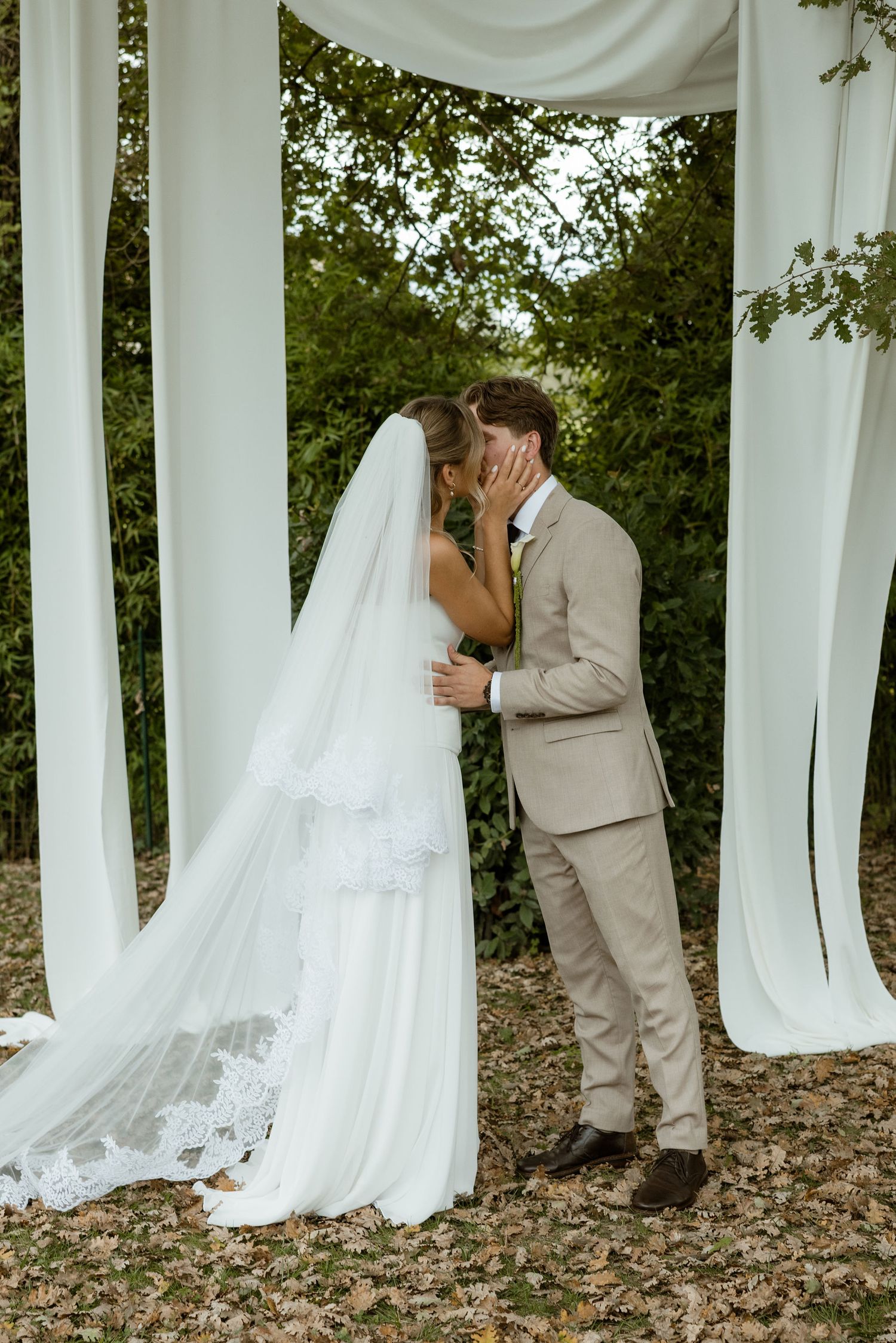 A romantic wedding moment captured under white fabric draping and trees with a bride in a flowing veil and groom in beige suit.