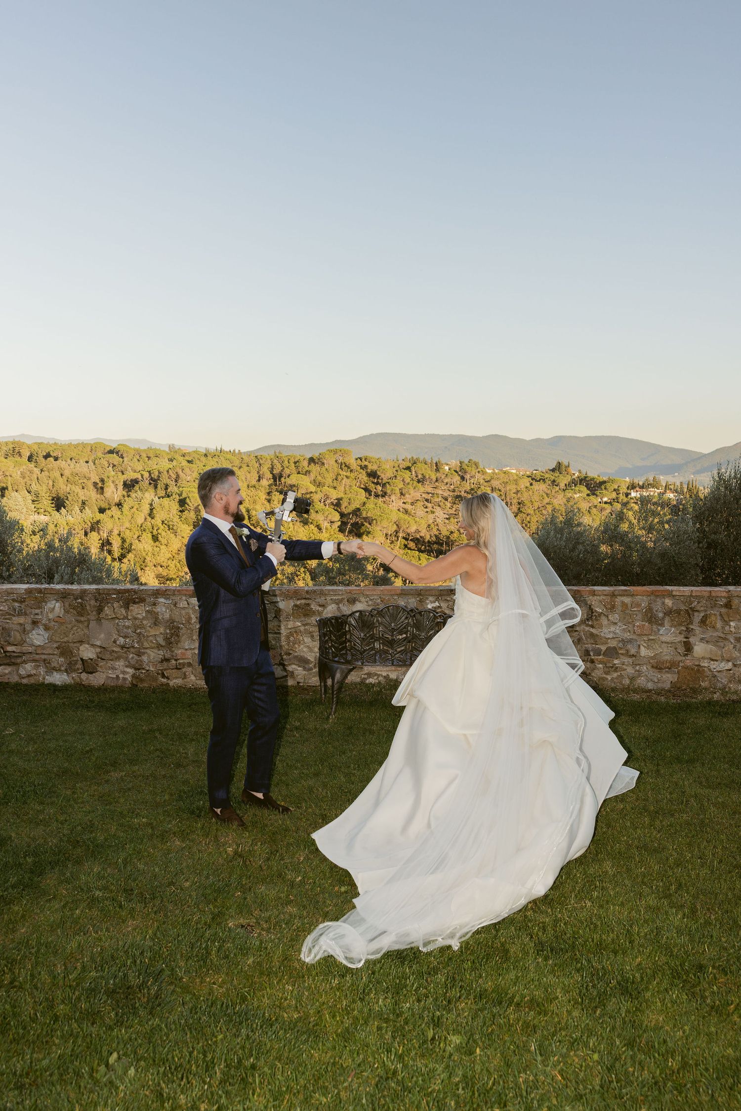 A bride and groom share a romantic moment on a grassy hilltop at sunset with mountains in the background.