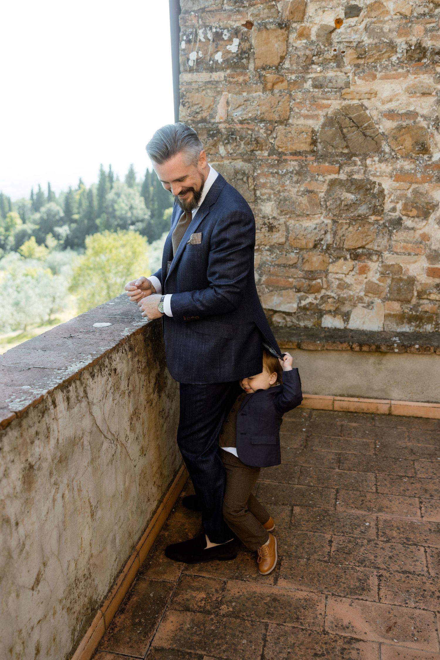 A person in a dark suit sits contemplatively on a stone ledge overlooking a scenic landscape.