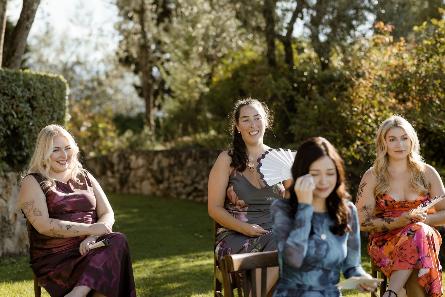 Four people seated outdoors enjoying a sunny day in a garden setting with manicured hedges.