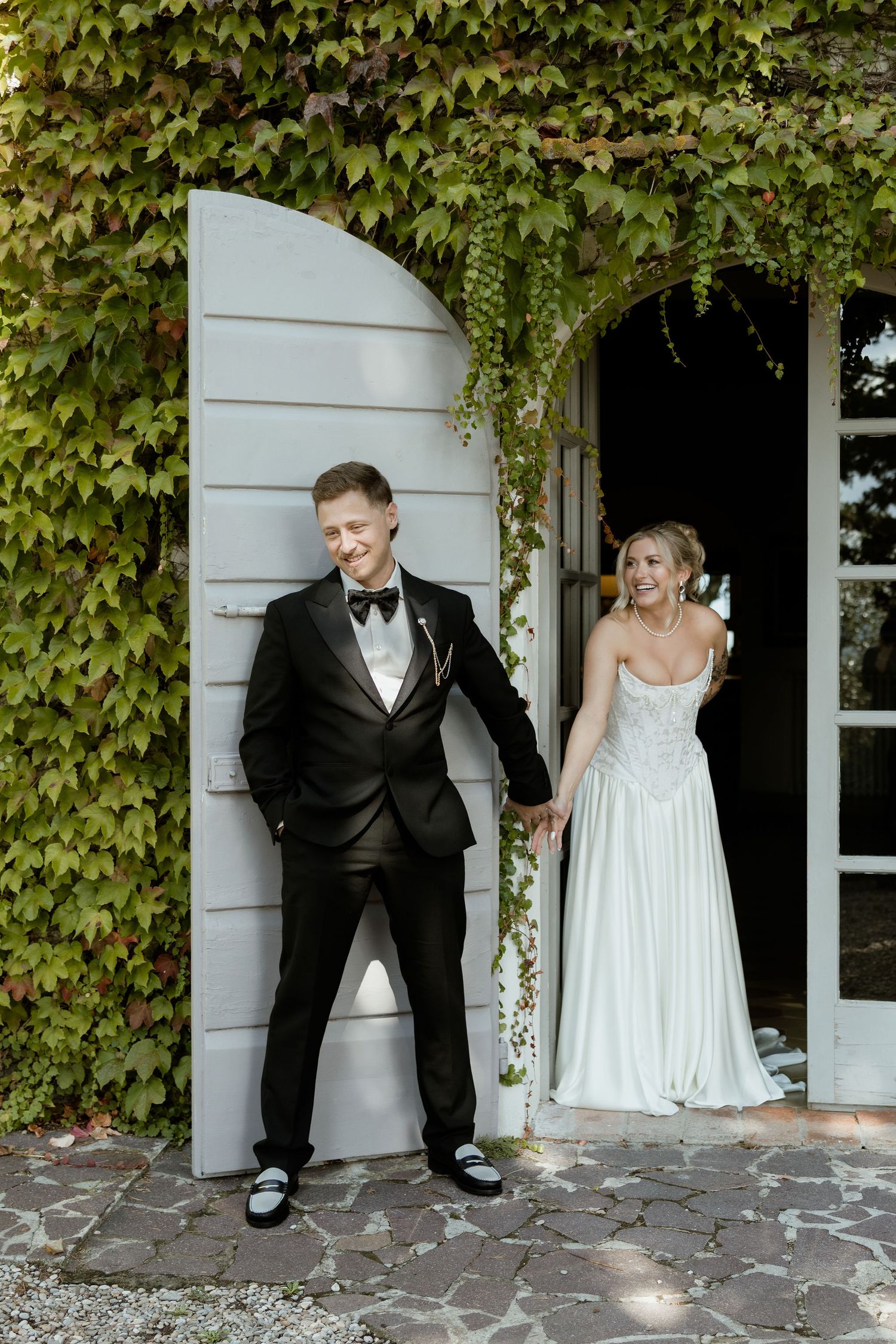 A playful wedding couple peeks out from behind white columns covered in lush green ivy vines.