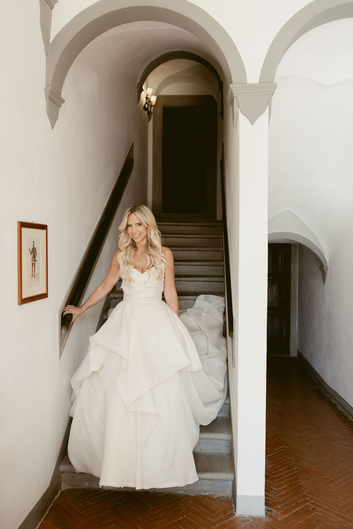 A bride in a flowing white wedding gown descends an elegant arched staircase in a historic building.