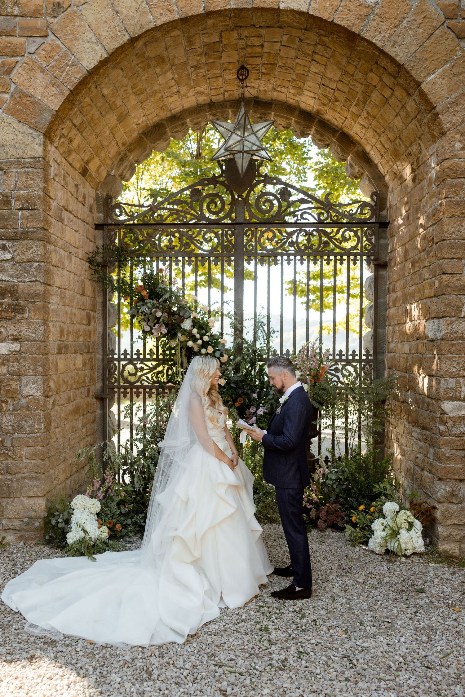 A wedding couple stands beneath a rustic stone archway with ornate iron gates and floral decorations.