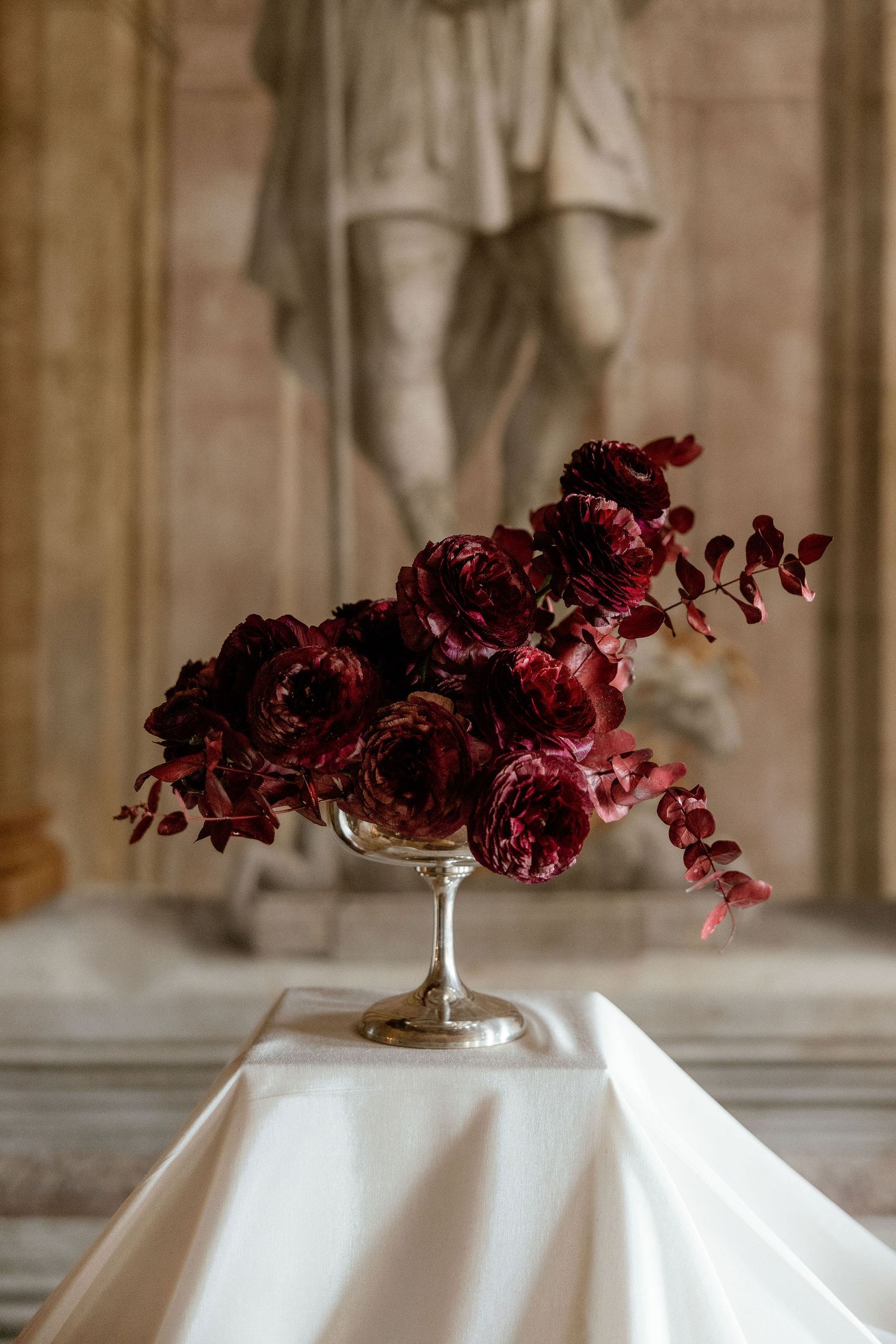 Deep burgundy roses and eucalyptus arranged in a silver compote vase on white linen against a stone backdrop.