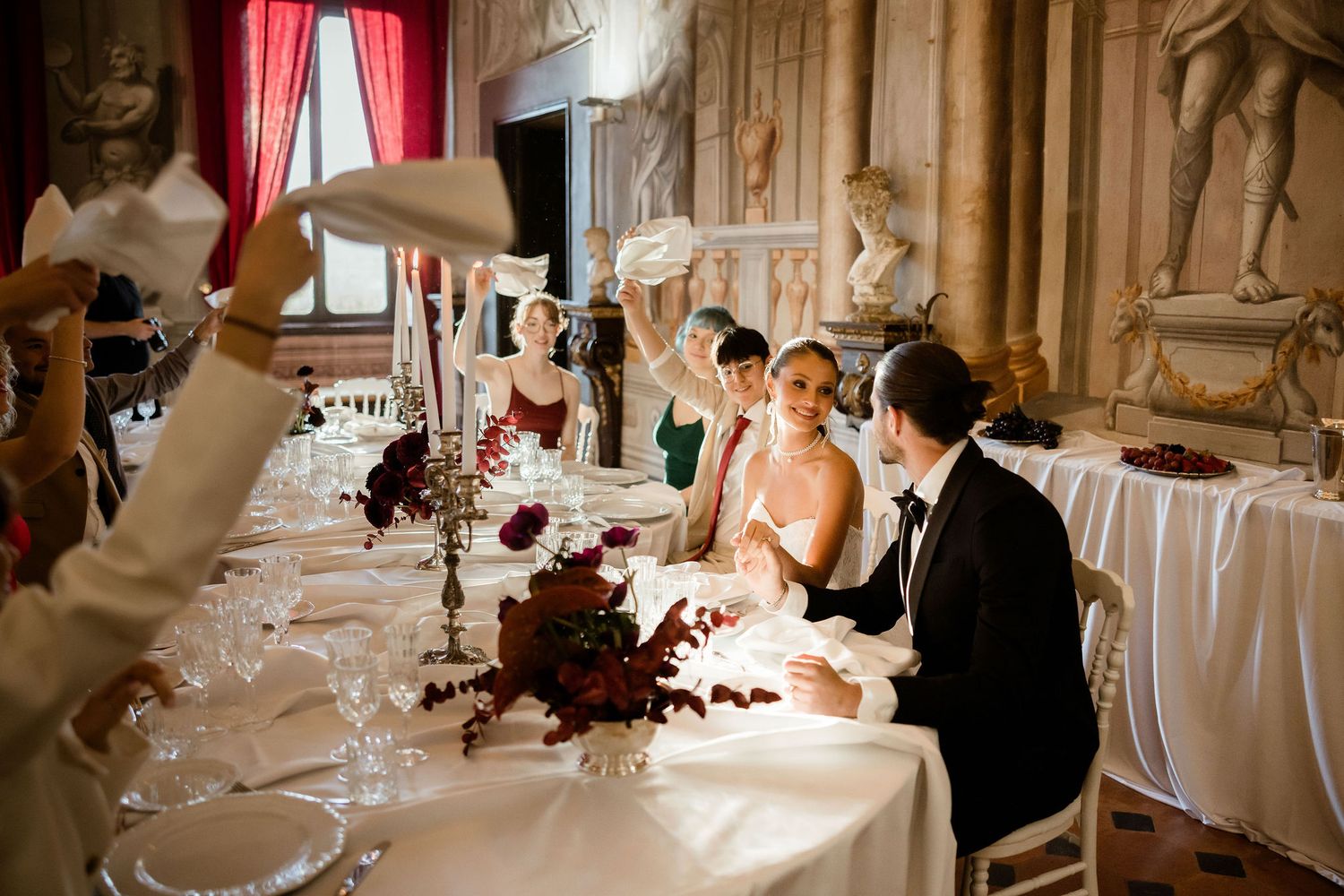 Wedding reception table setup in an ornate baroque hall with decorative statues and burgundy floral centerpieces.