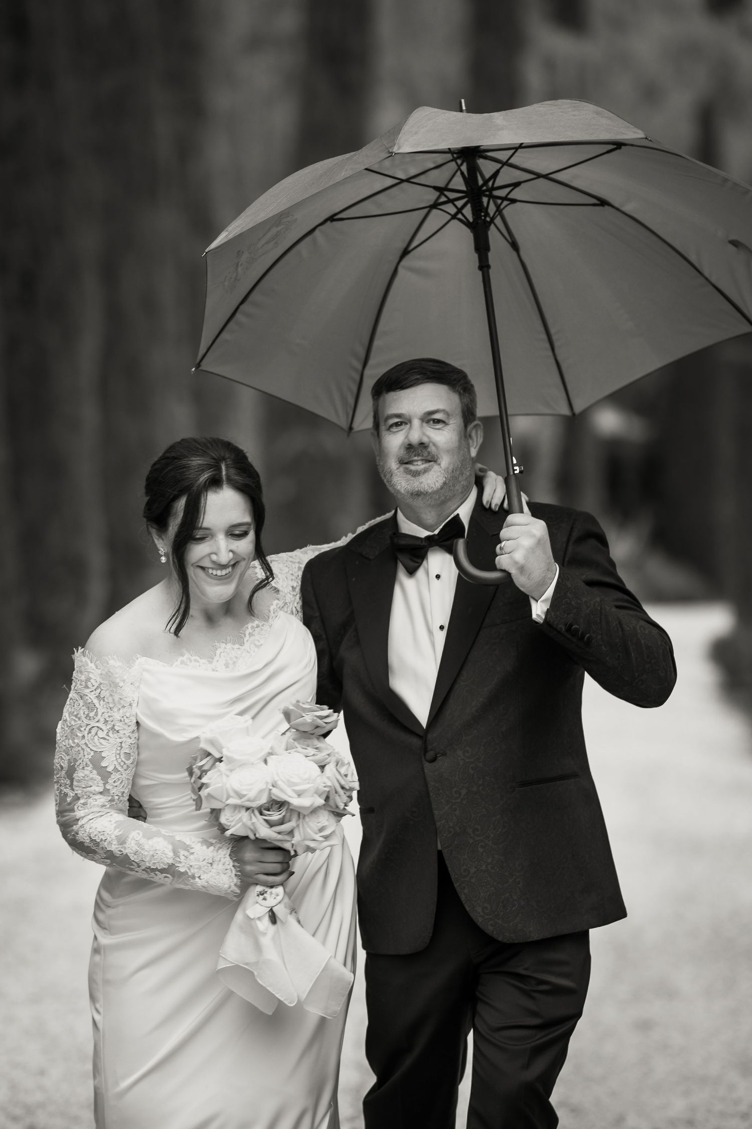 Black and white wedding photo with couple walking together under white umbrella in formal attire with bridal bouquet.