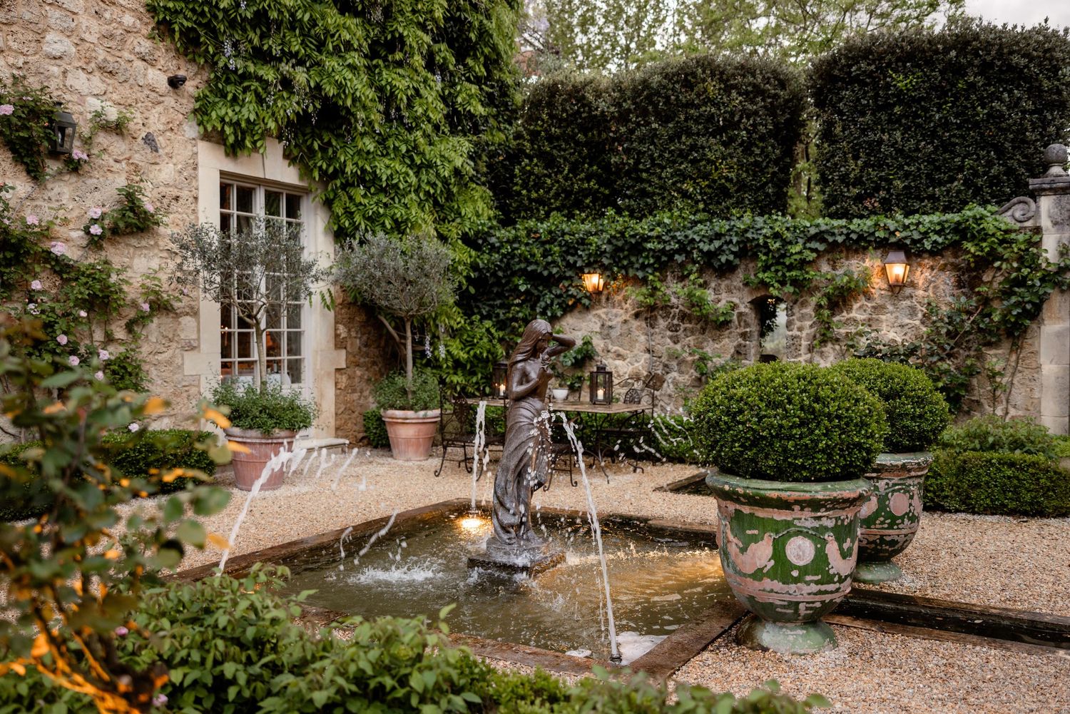 A charming garden courtyard features stone walls covered in ivy, a central fountain, and classic garden urns with topiary.
