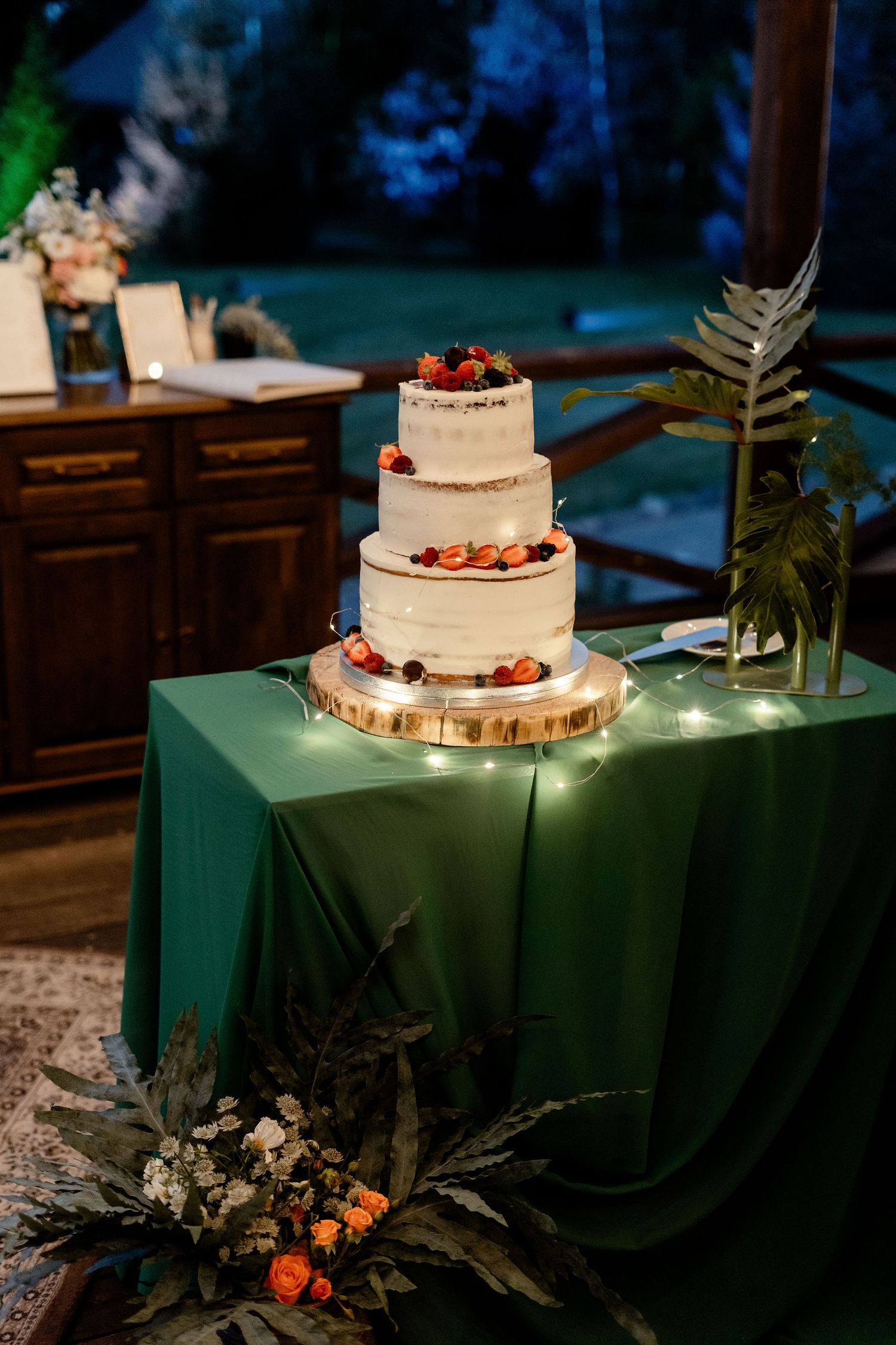 A three-tier white wedding cake with red berries sits on a wooden stand covered by an emerald green tablecloth.