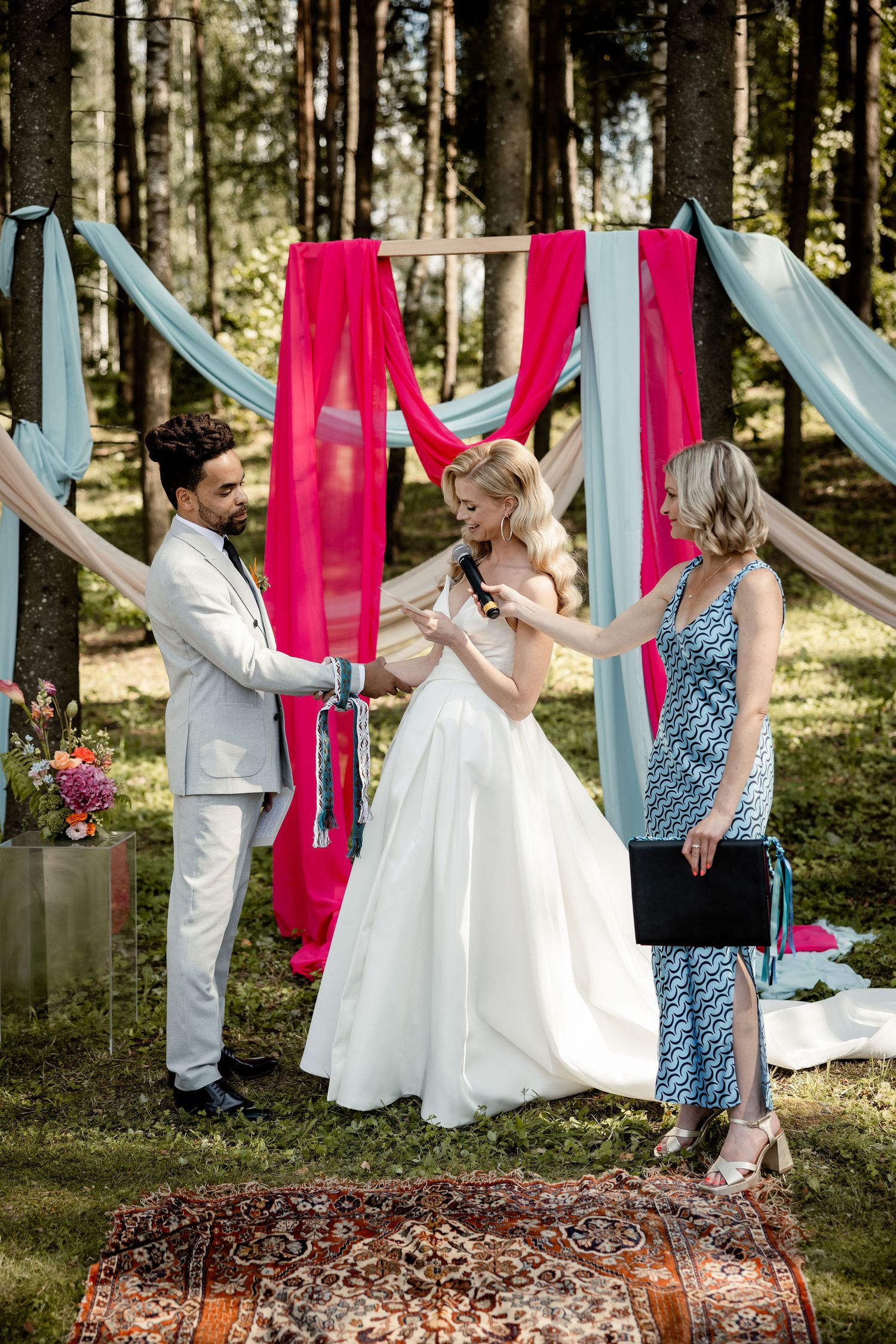 A wedding ceremony in a forest with hanging red and blue fabric decorations and a vintage rug on the ground.