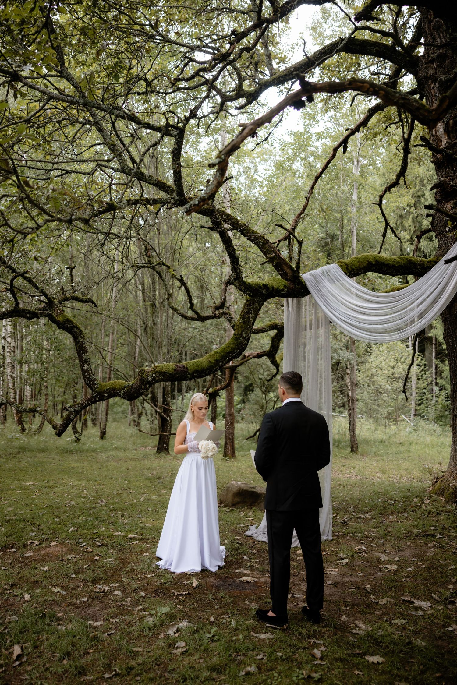 A couple stands beneath a rustic wooden arch with white draping and mossy tree branches in a woodland setting.