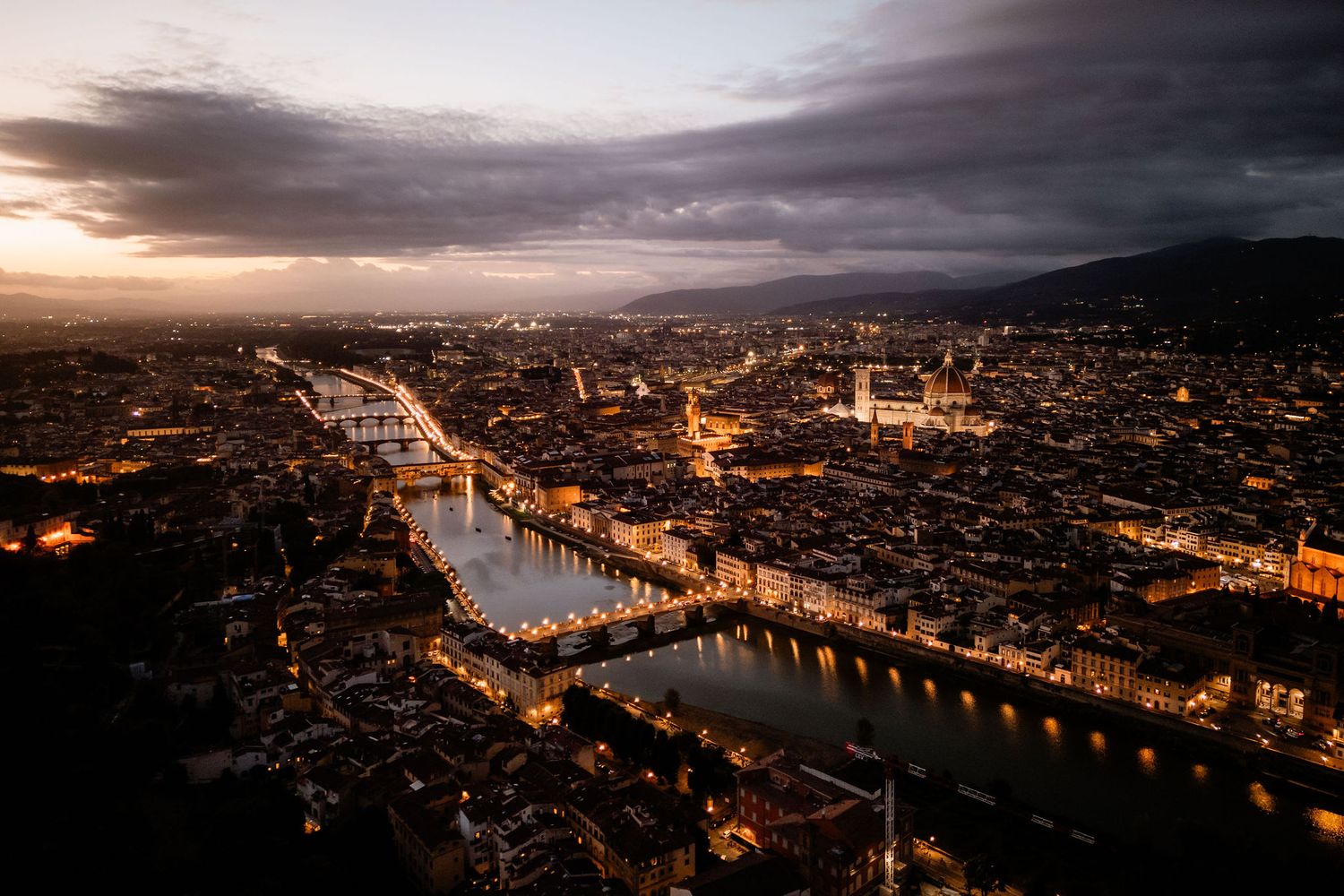Aerial cityscape of Florence at sunset with illuminated bridges crossing the Arno River and historic buildings glowing.