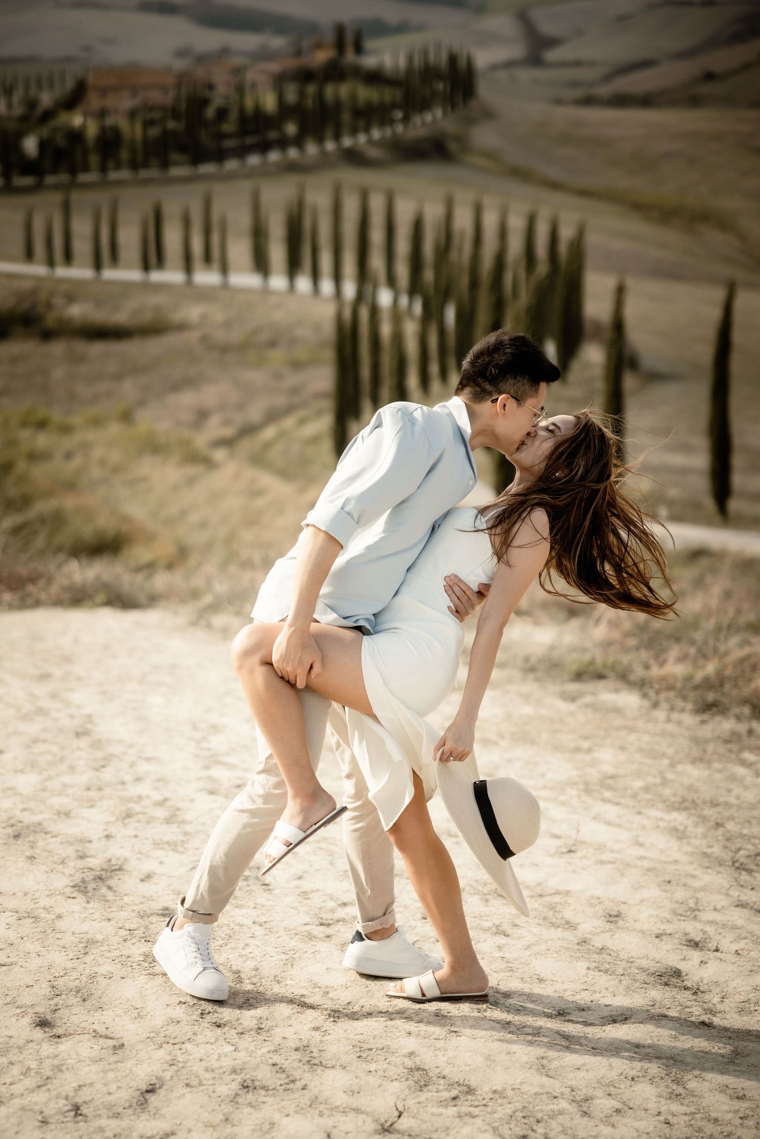 Dancing couple in white outfits sharing romantic moment against Tuscan cypress landscape.