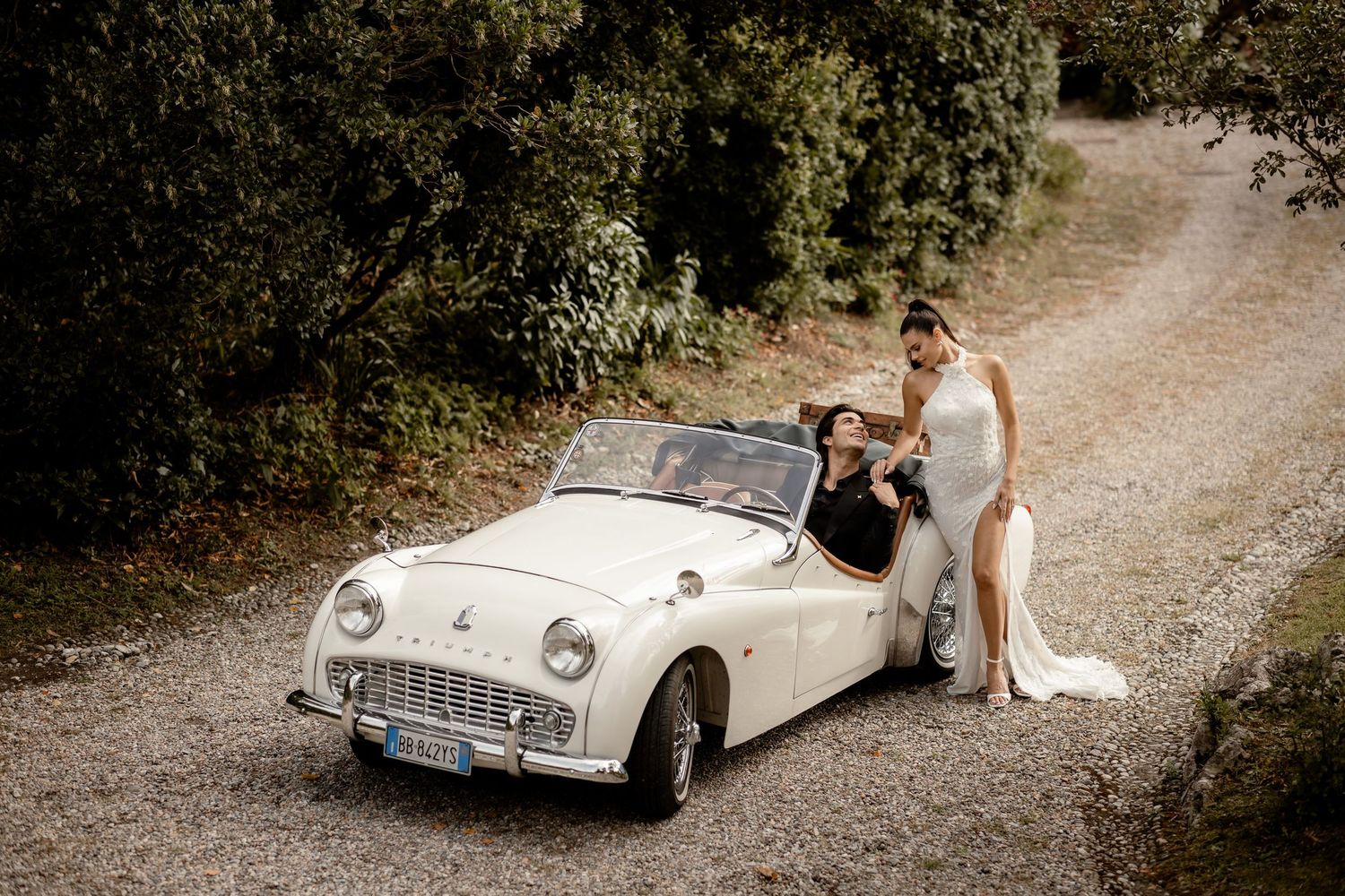 Newlyweds pose with classic white Triumph car on country road.