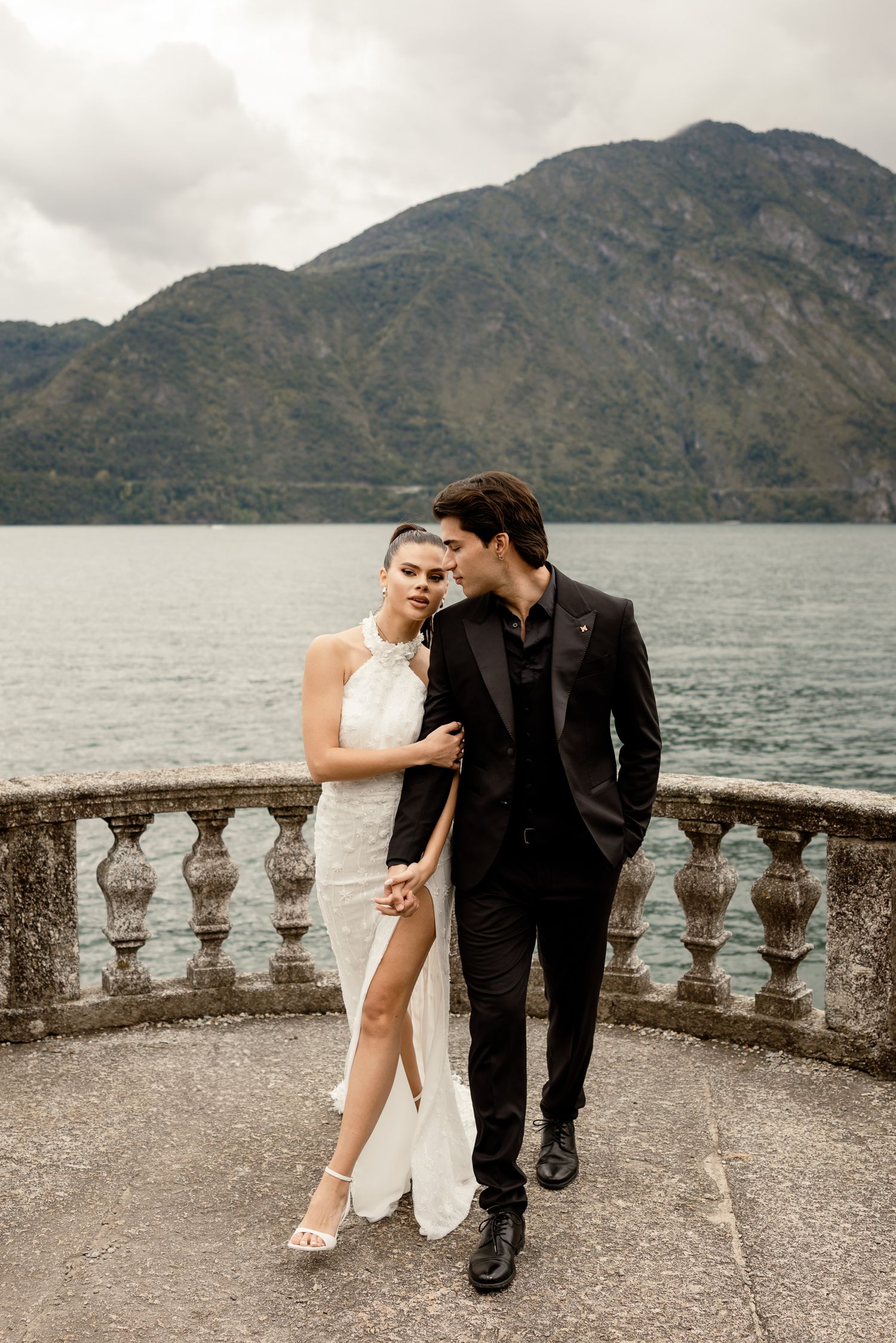 Couple in formal attire embrace on a stone balcony overlooking a mountain lake.