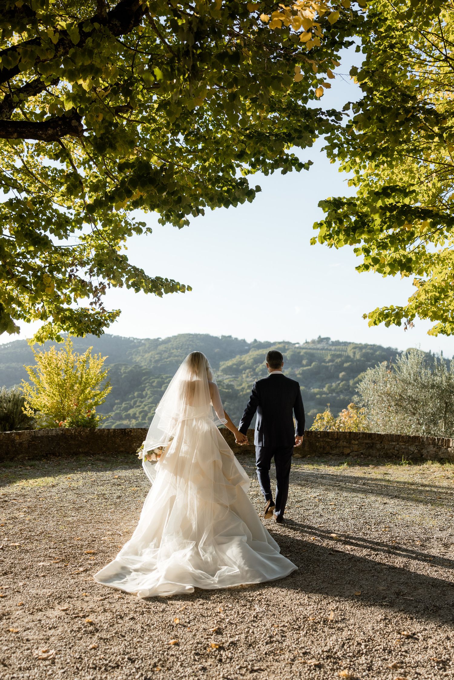 A bride and groom walk hand in hand on a mountain path surrounded by autumn trees and scenic views.