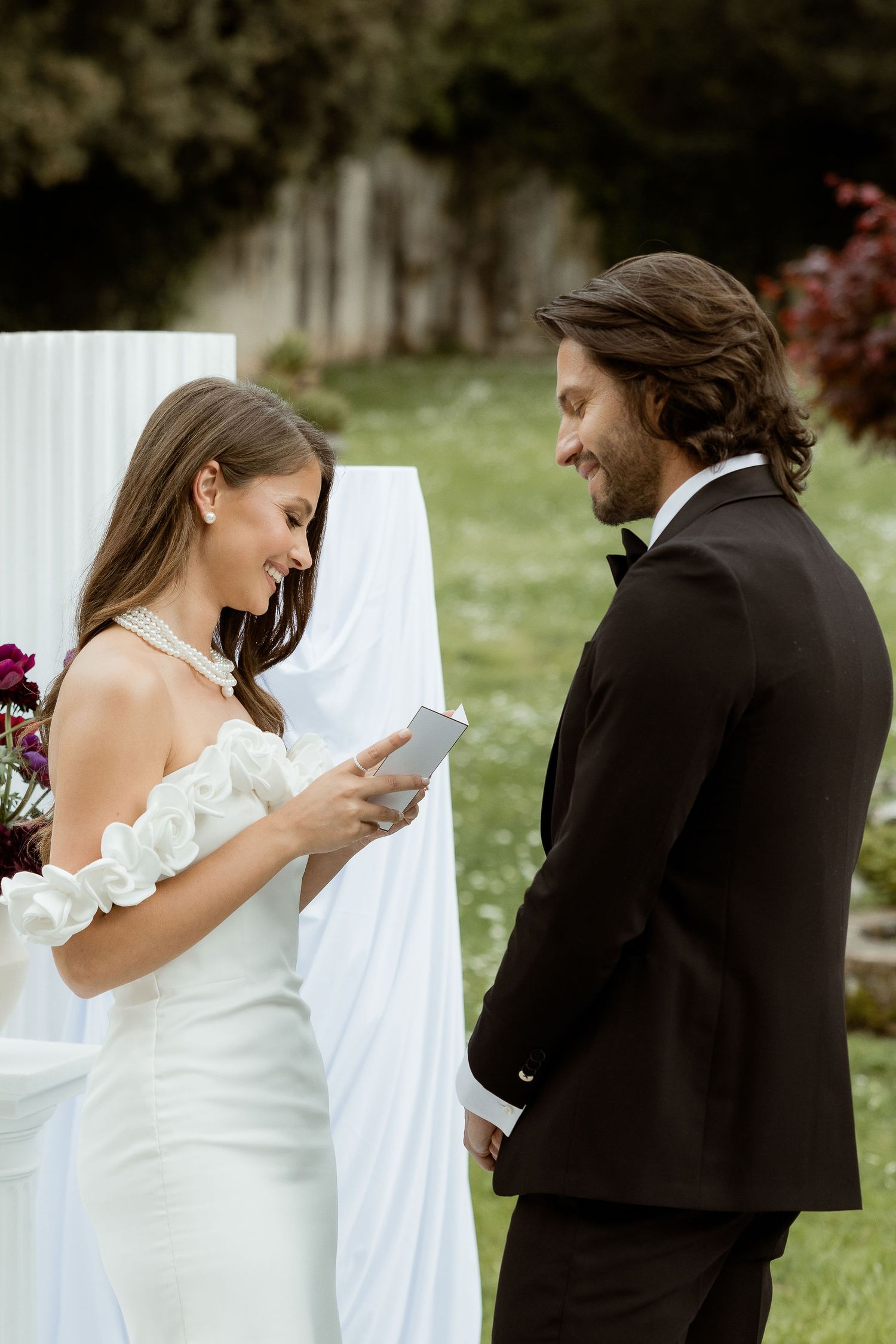 A couple reading their wedding vows during an intimate outdoor ceremony with white drapery and flowers in the background.