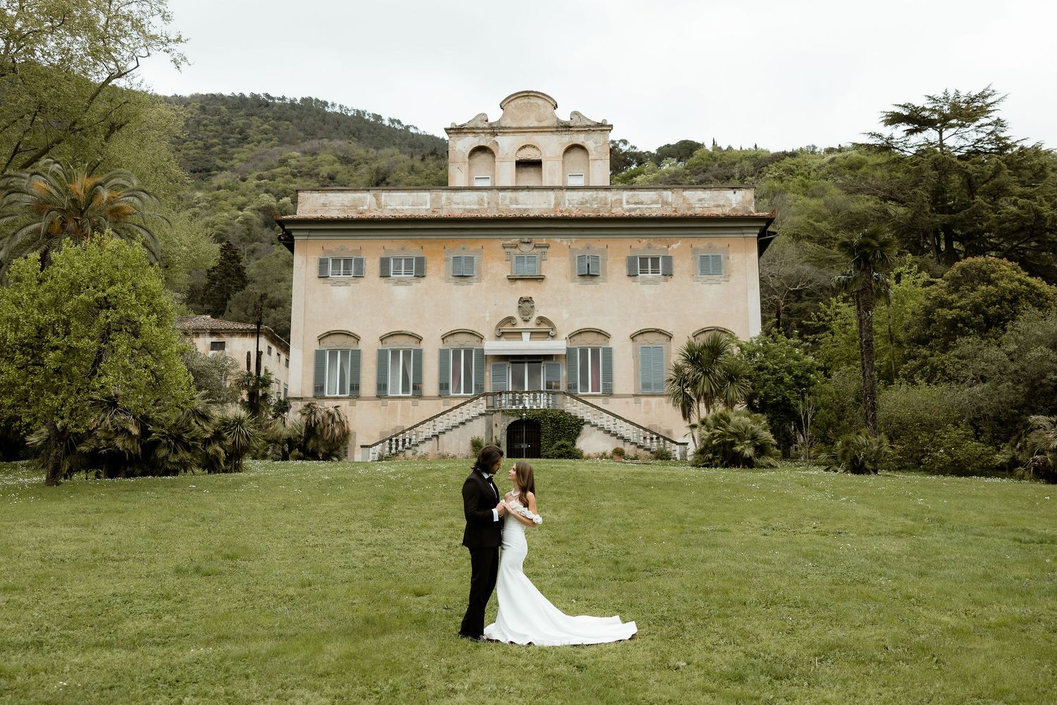 Elegant villa with cream facade and grand staircase serves as backdrop for wedding couple on manicured lawn.