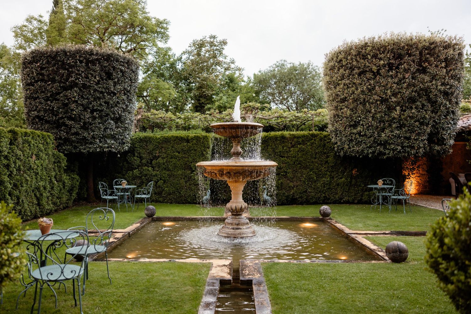 Elegant garden fountain centerpiece surrounded by manicured hedges and turquoise patio chairs in a formal garden setting.