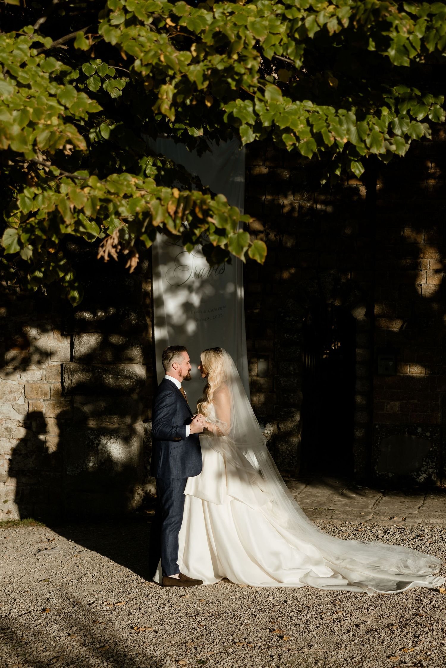 A romantic wedding couple shares an intimate moment under the shade of trees on a gravel path.