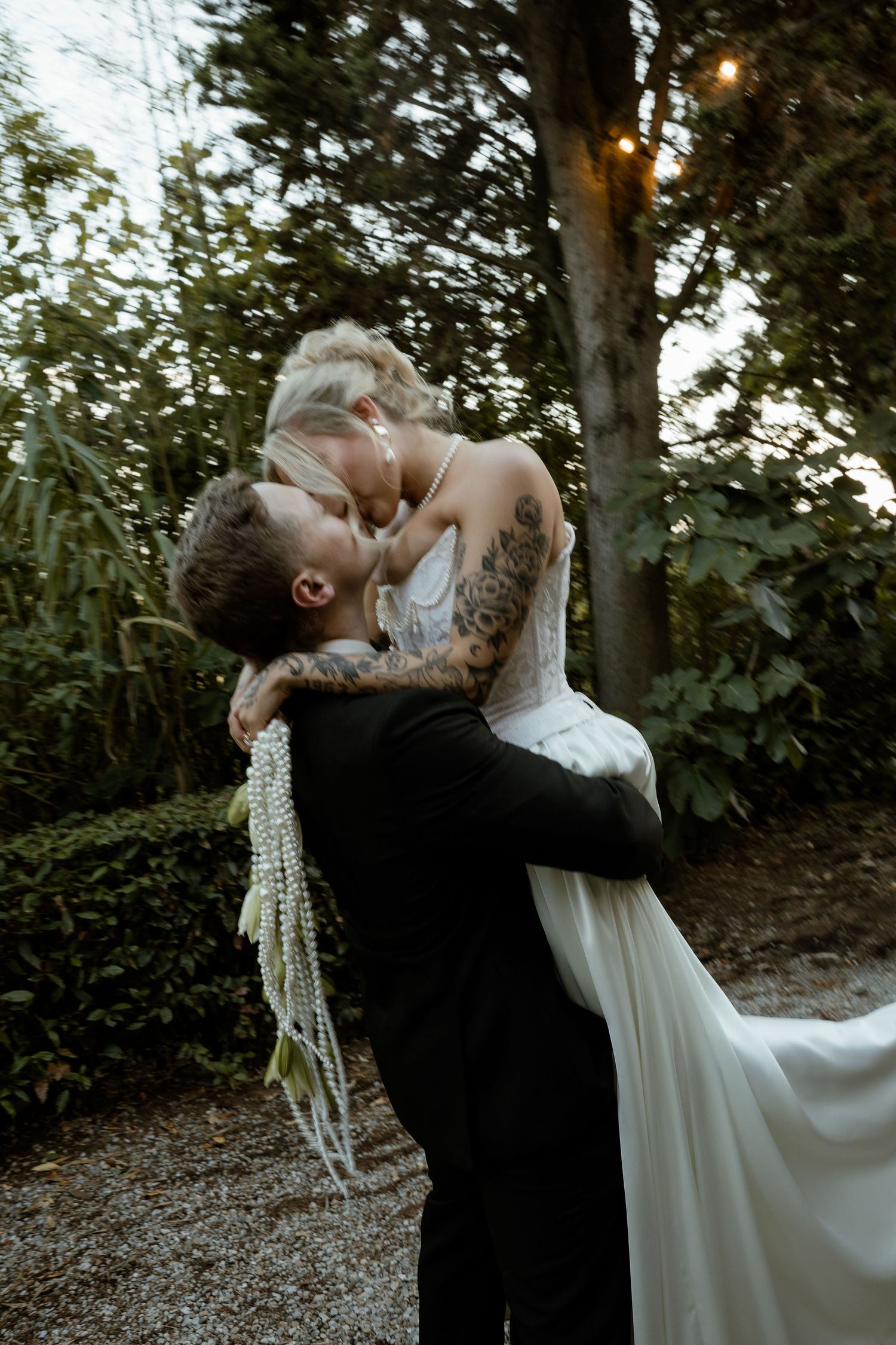 A romantic wedding couple shares an intimate dip kiss in a wooded outdoor setting at dusk.