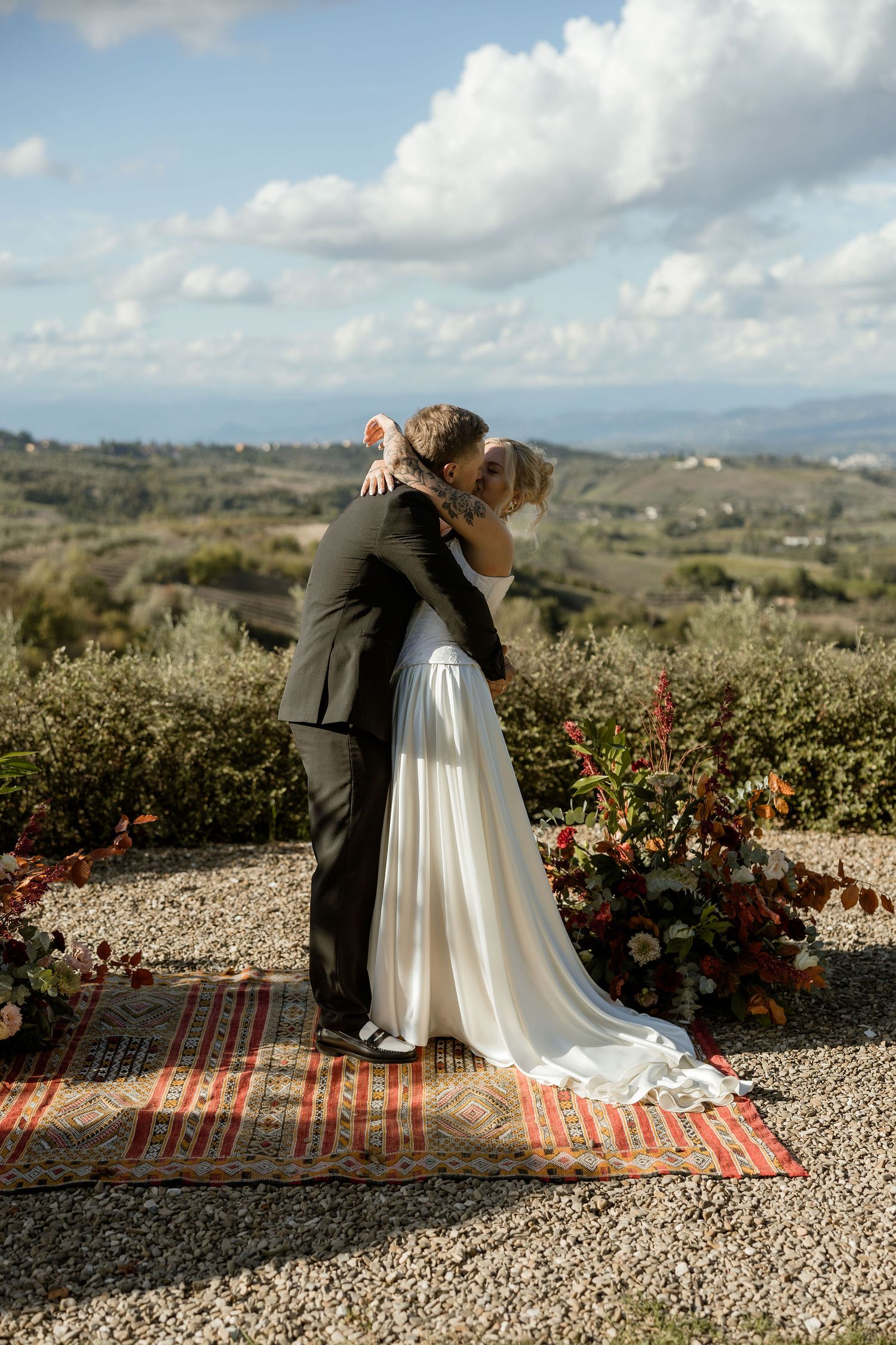 A couple in wedding attire embrace on a gravel path overlooking rolling hills and countryside on a cloudy day.