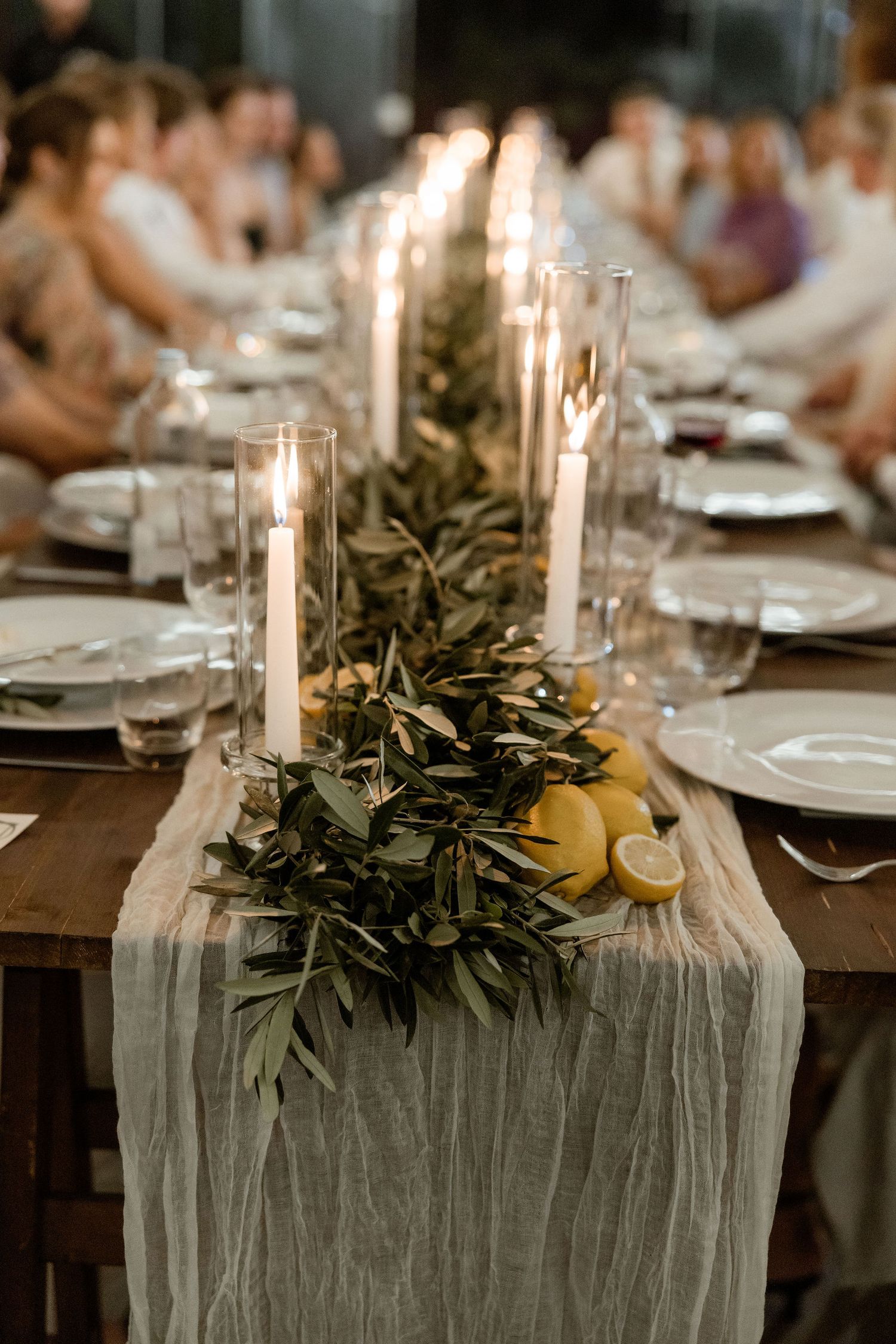 Elegant rustic table setting with white candles, lemon slices, and olive branch garland on gray linen runner.