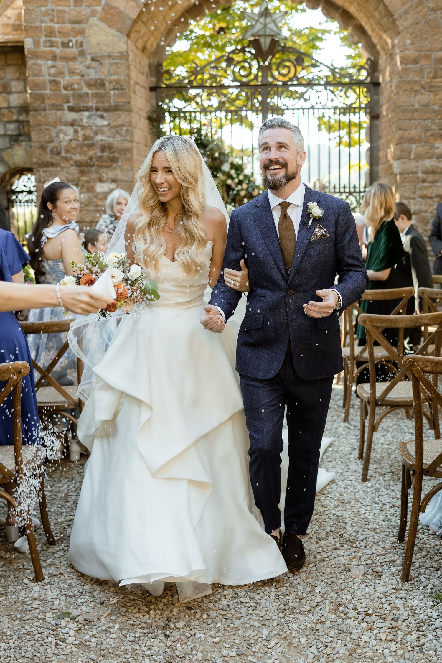 Wedding guests throw rice and confetti at newlyweds under rustic stone archways during outdoor celebration.