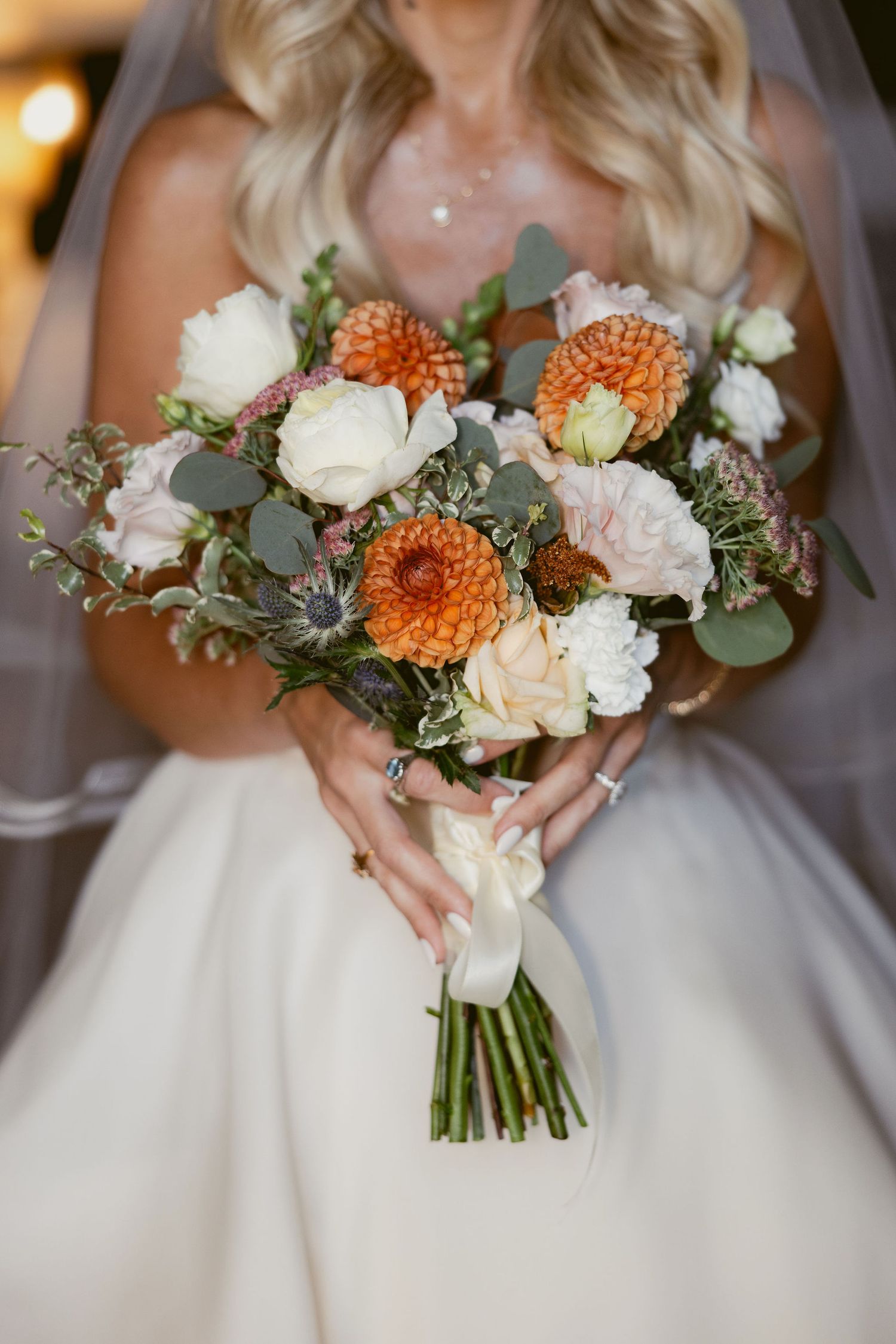 A series of wedding bouquets featuring white roses, orange dahlias, and eucalyptus greenery held against white gowns.