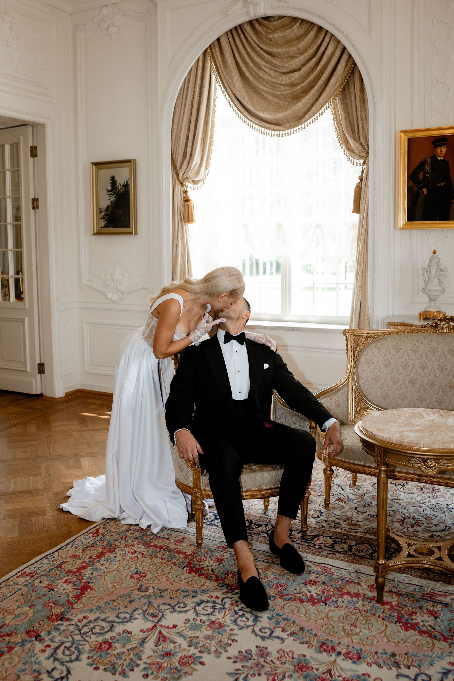 Couple in wedding attire sharing a tender moment by an ornate window with flowing drapes.