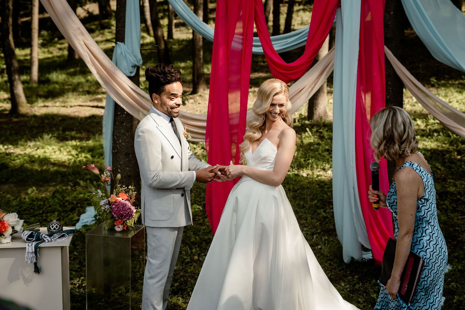Couple exchanges vows during woodland ceremony with vibrant red and blue draped fabric decor.