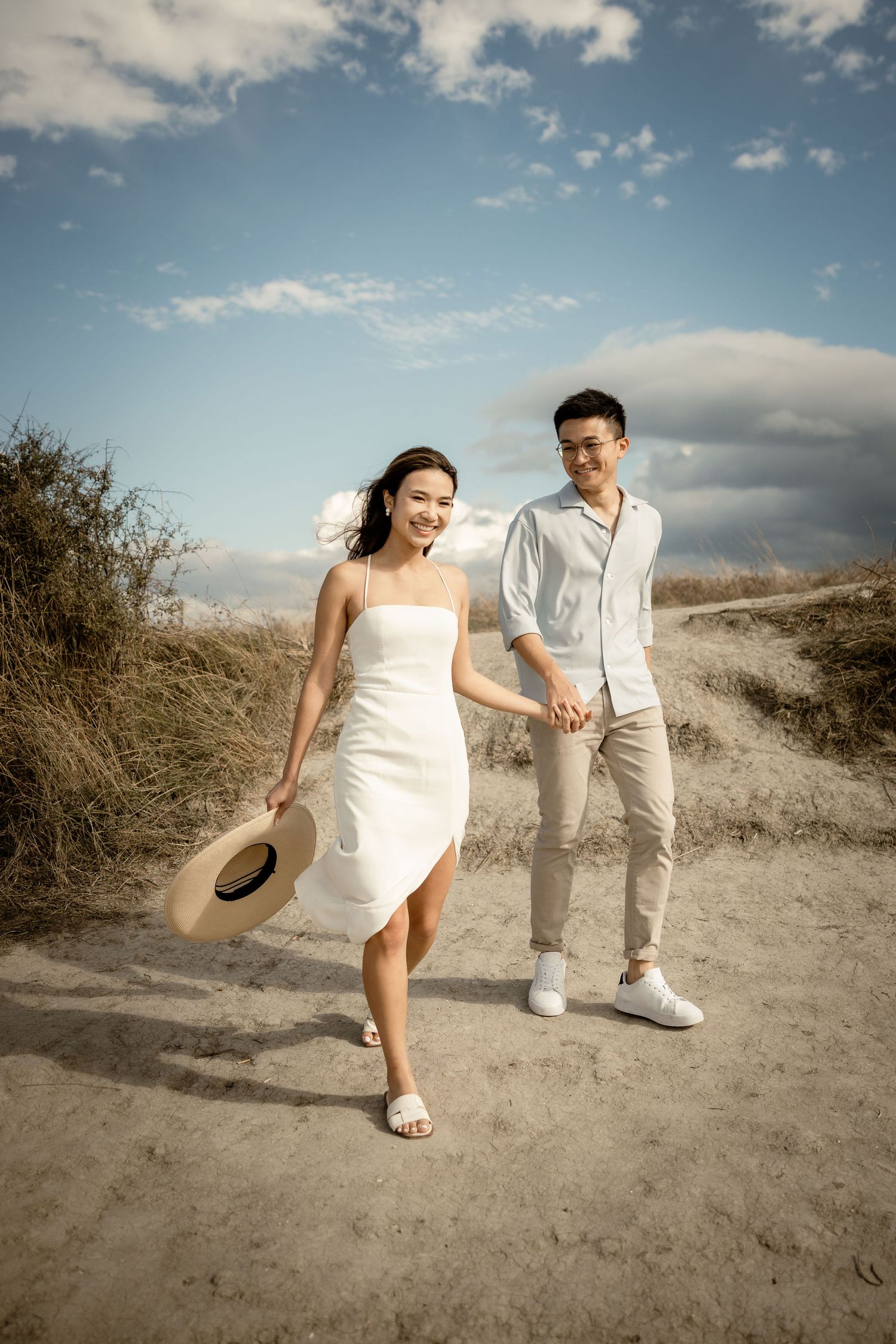 Couple in white outfits strolling along a sandy trail with a straw hat.