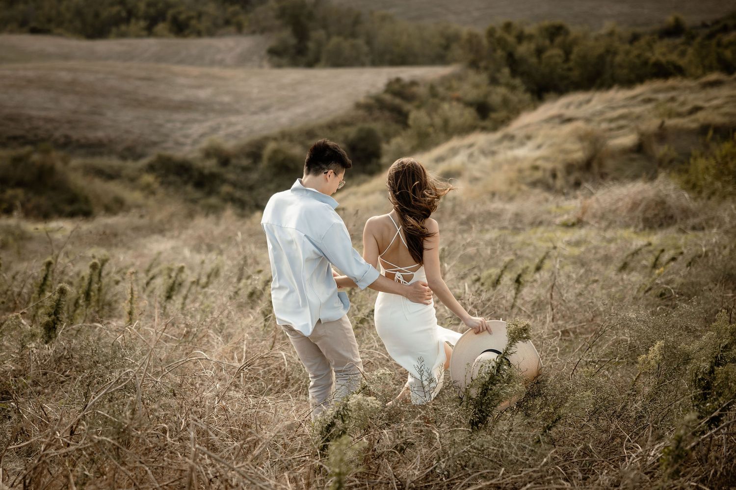 A couple descends a winding path through golden grass on a hillside at sunset.