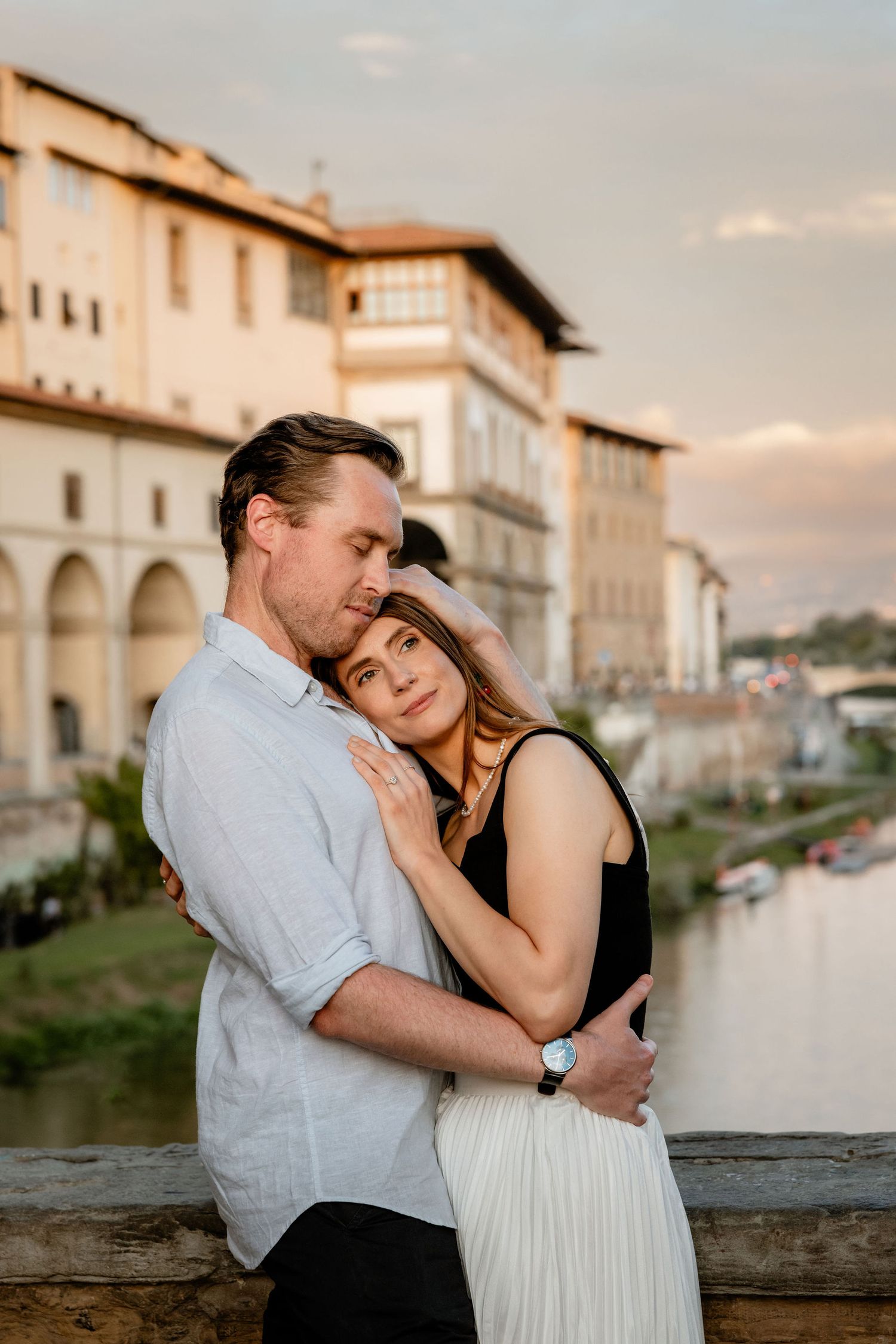A couple embracing by a scenic Italian river with historic architecture in the background during sunset.