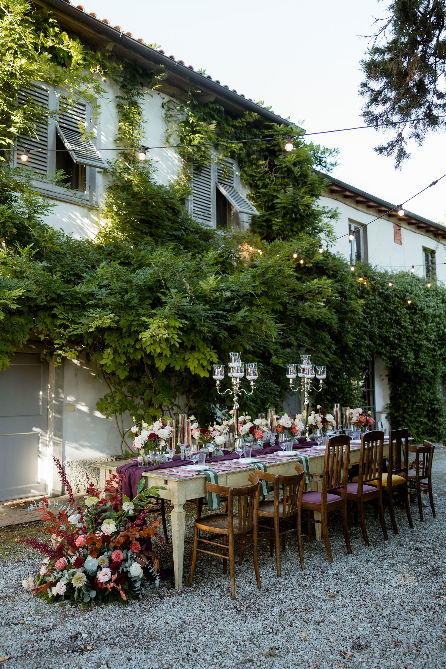 Long outdoor dining table setup with rustic chairs under lush green vines and floral arrangements on gravel ground.