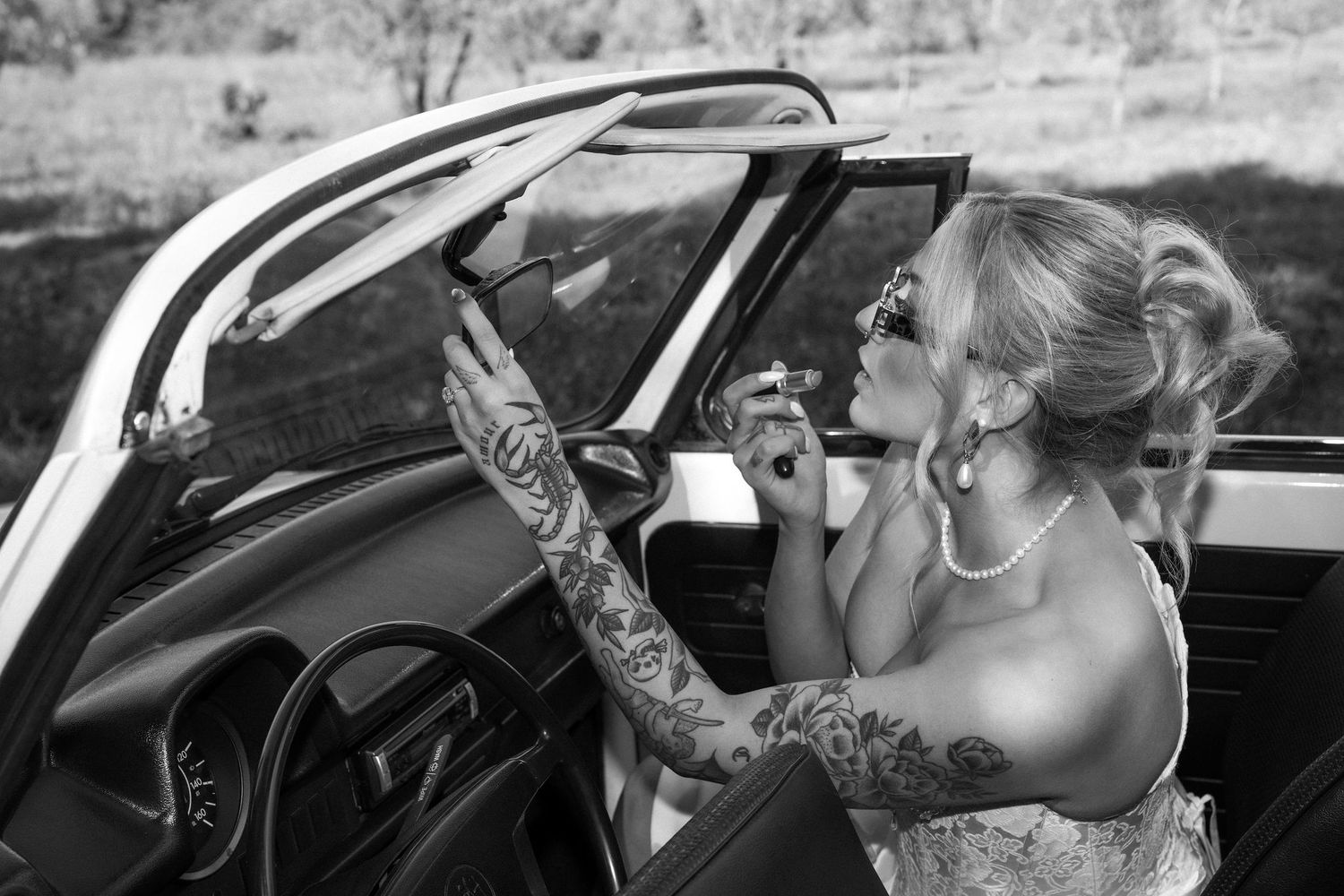 A woman applies lipstick while looking in a car mirror, showing detailed tattoo sleeve in black and white photography.