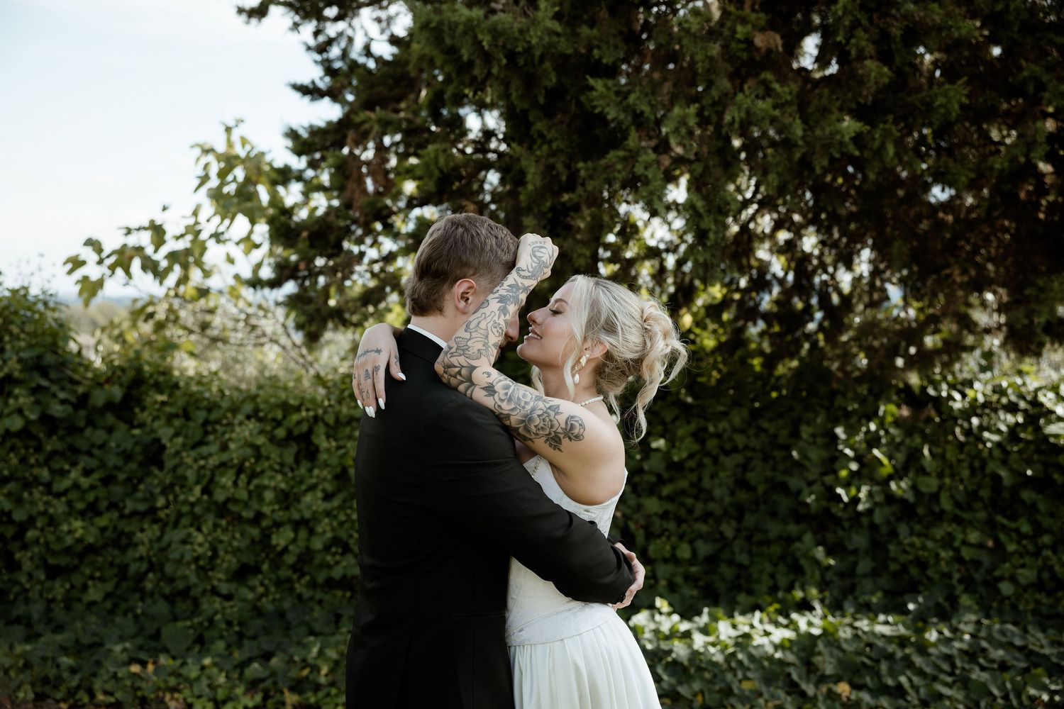 A couple in wedding attire embraces while dancing outdoors under trees and string lights at dusk.