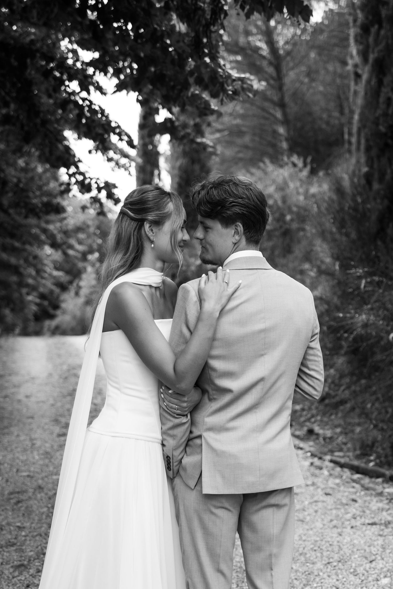 A romantic black and white photo series of a couple walking together on a gravel path surrounded by trees.
