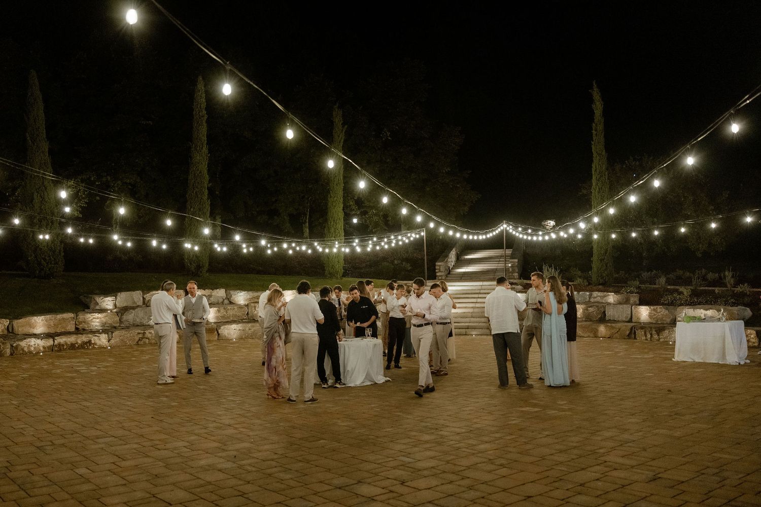 String lights illuminate an outdoor beach wedding reception at night with guests mingling on the sandy shore.