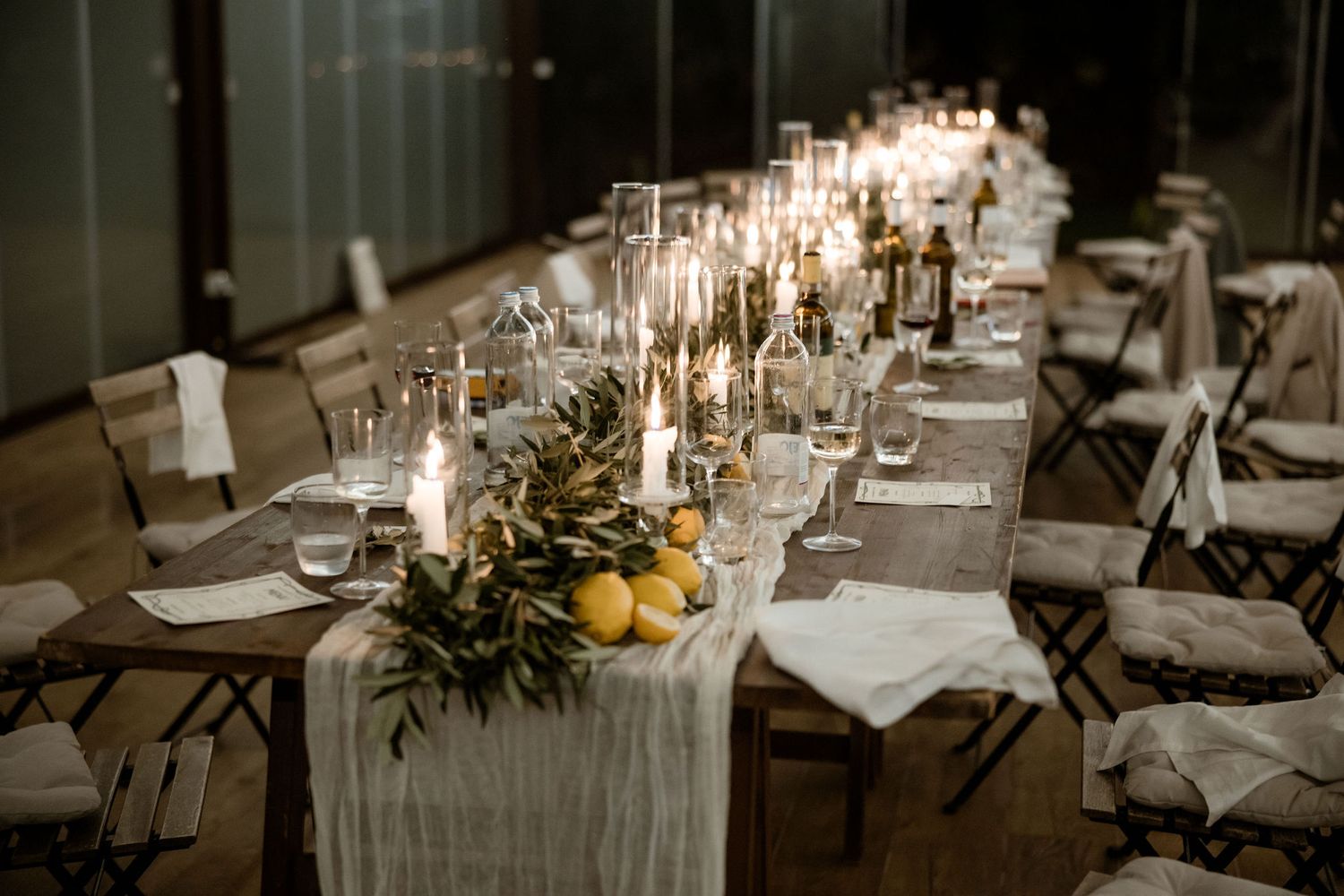 Long dining tables decorated with white linens, candles, and greenery create an elegant wedding reception atmosphere.