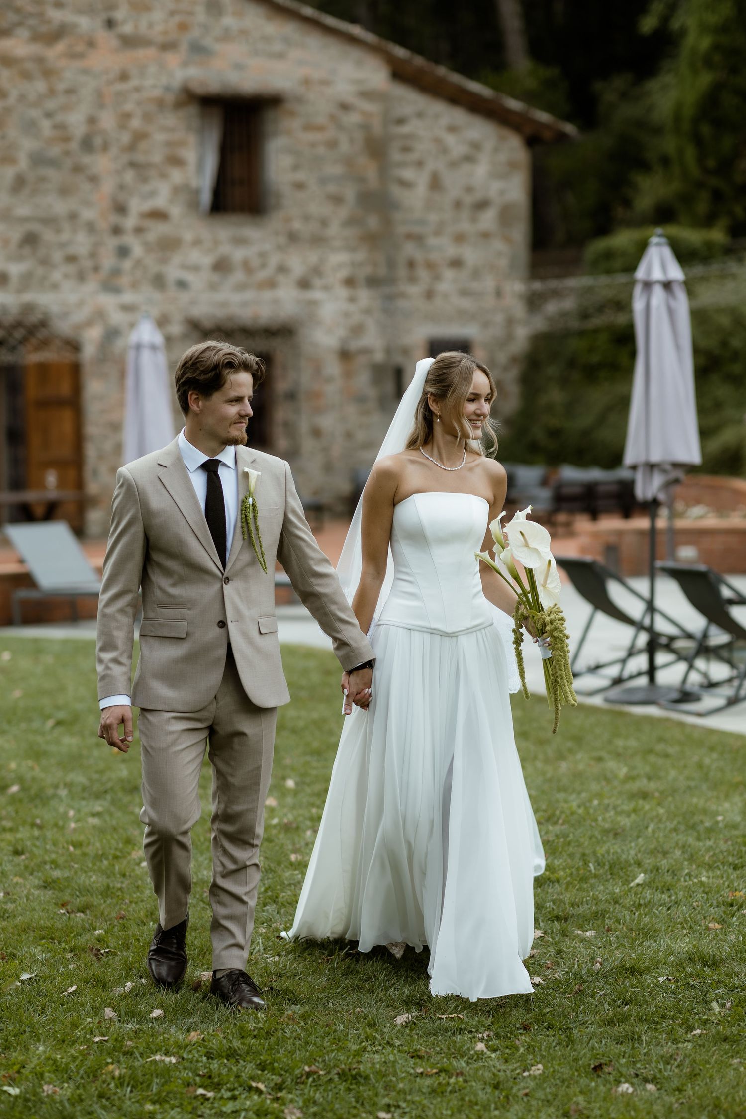 A wedding couple walks together on a grassy lawn outside a rustic stone building with white umbrellas in the background.