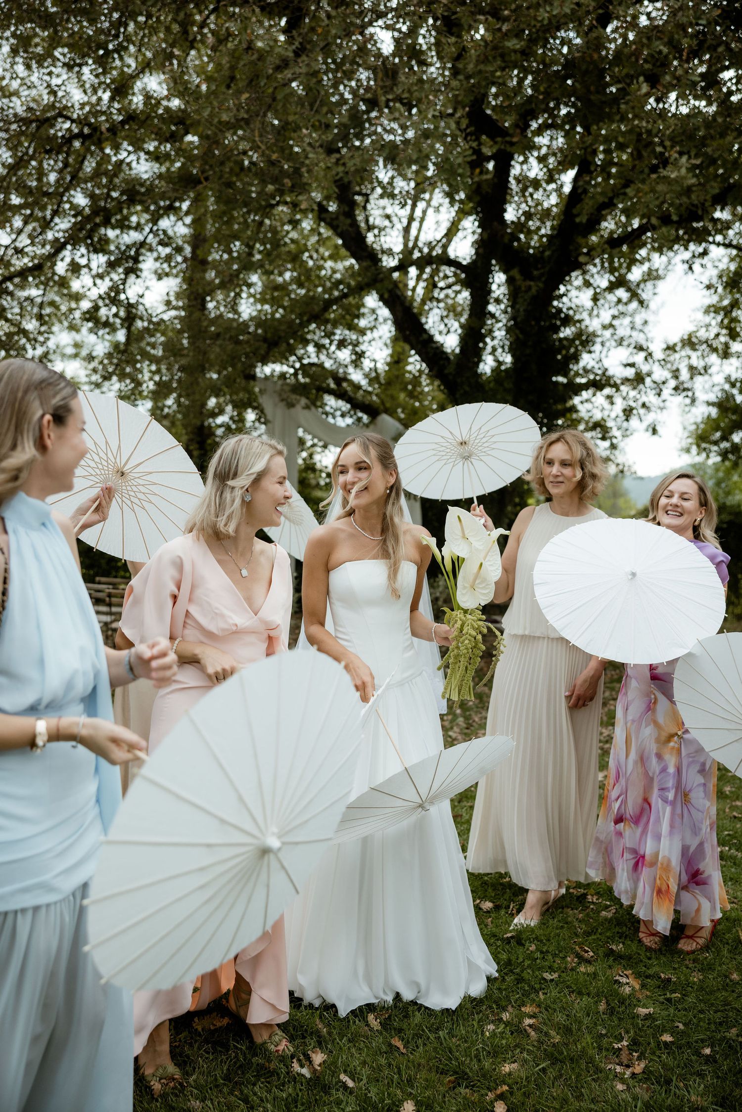 Group of women in light colored dresses twirl with white paper parasols during an outdoor photoshoot.