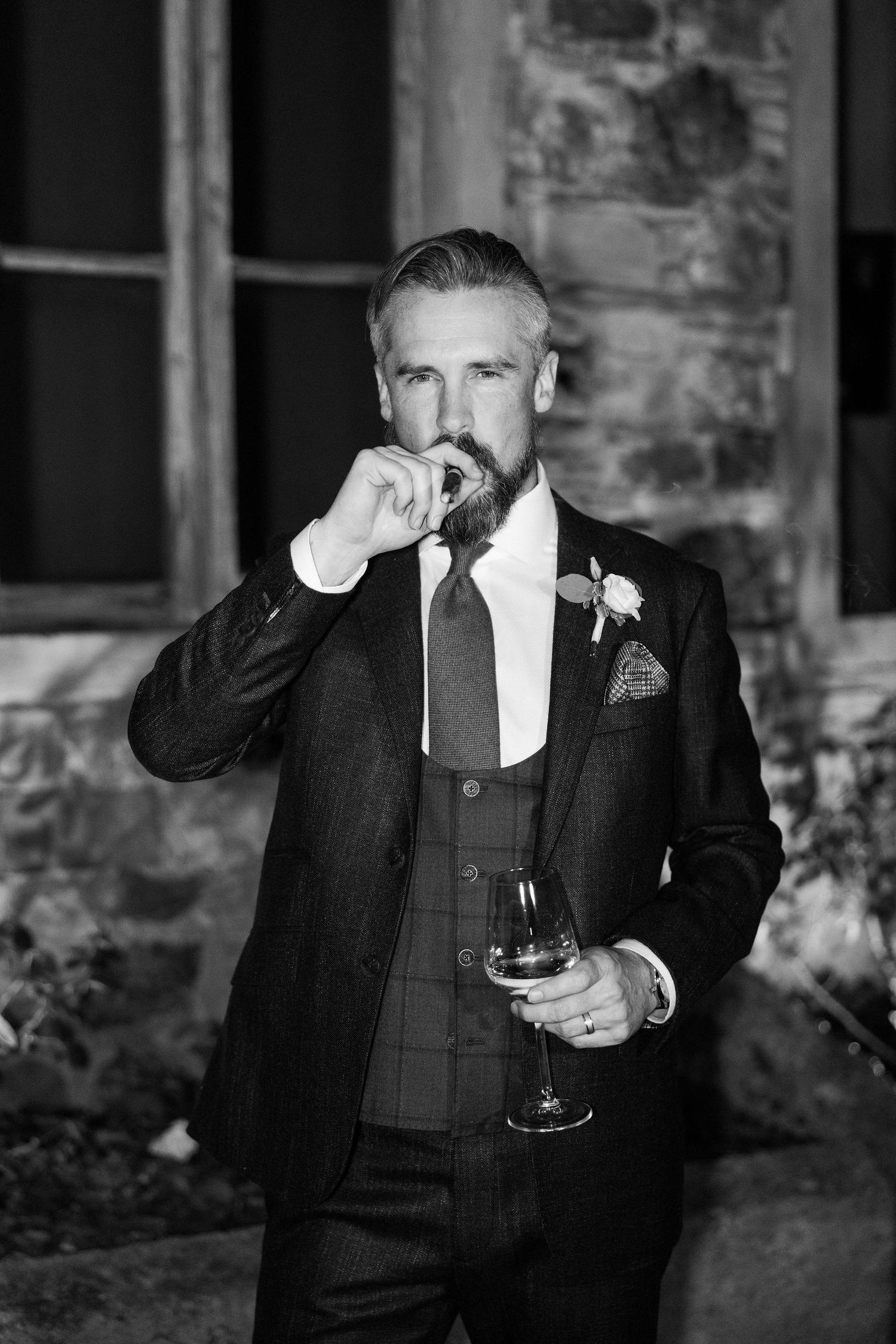 A formal portrait of a well-dressed gentleman in a dark suit enjoying a cigar and wine against a stone wall.