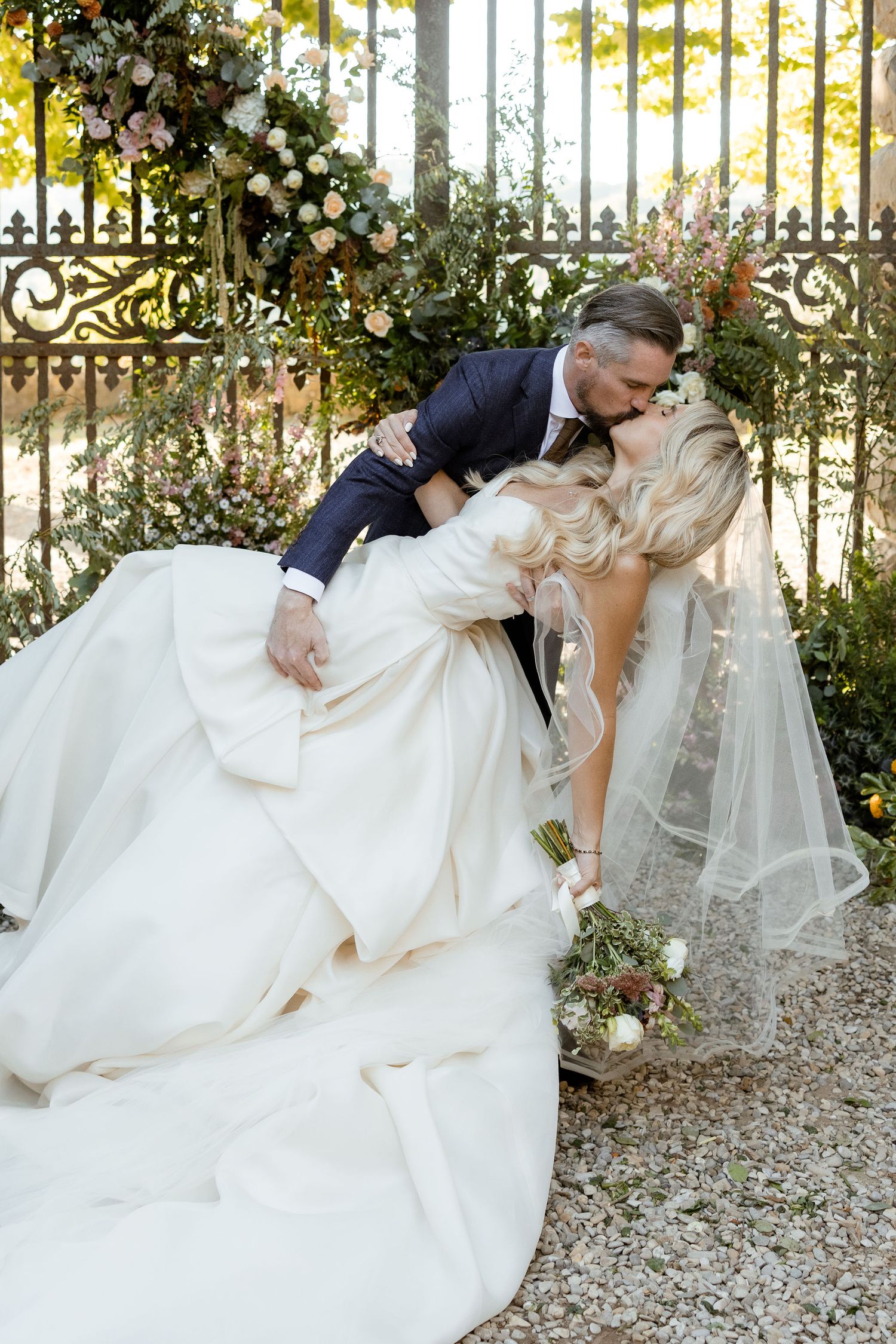 Romantic wedding couple in traditional attire share intimate dips in a garden setting with white gravel and iron gates.