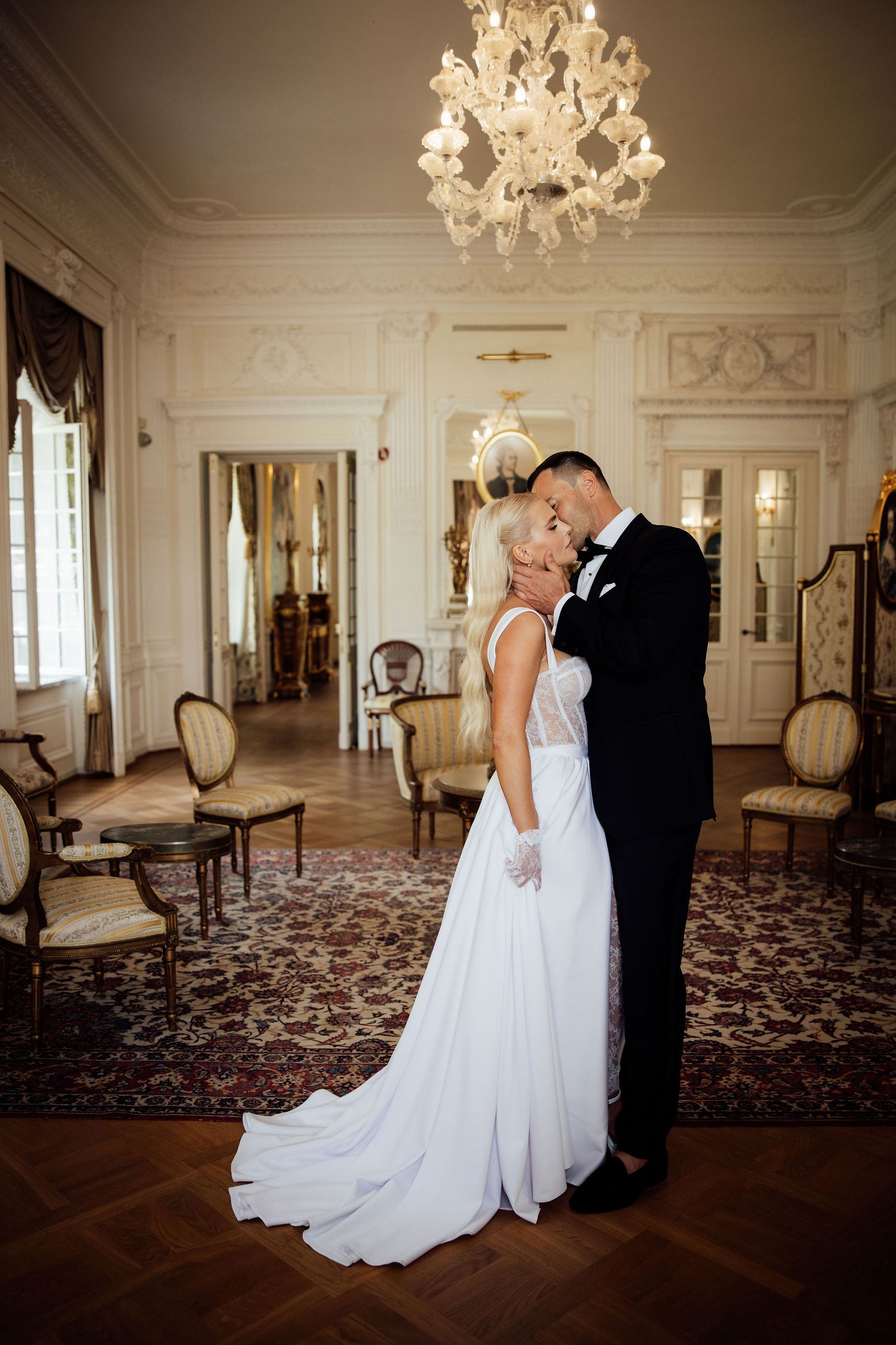 A bride in white gown and groom in tuxedo share an intimate moment beneath an ornate crystal chandelier.